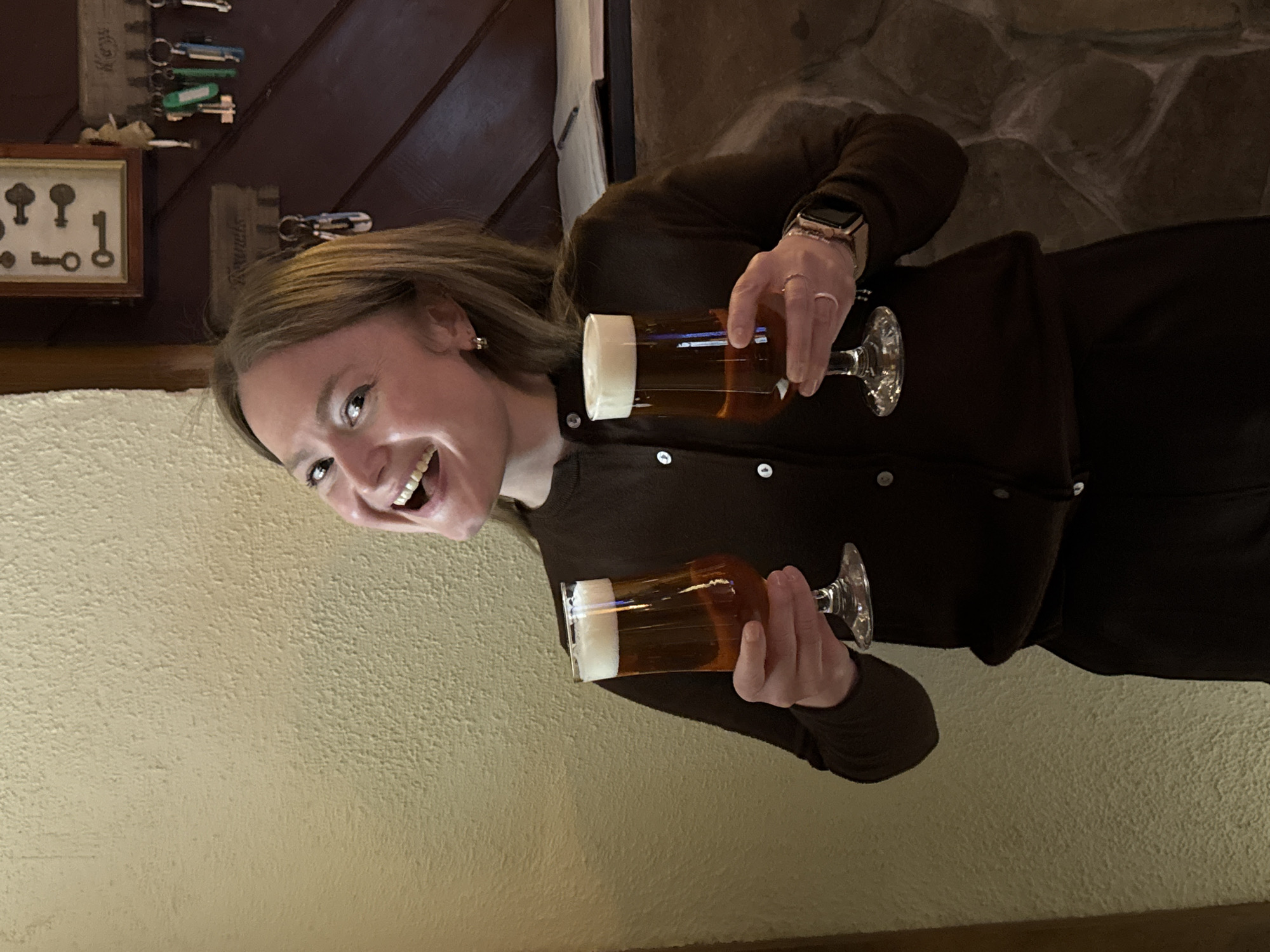A female subject, appearing adult and Caucasian, with light brown hair, is positioned centrally, facing forward, and smiling broadly with an open mouth. She wears a dark brown long-sleeved cardigan with white buttons and dark lower garments. On her left wrist, a rose gold-colored smartwatch is visible, along with a ring on her left ring finger. Small hoop earrings are worn in both ears. She holds a stemmed glass containing an amber liquid topped with a white foam head in each hand, presenting them towards the viewer. The background to the left features a light yellow, textured wall. To the right, dark brown wooden paneling with diagonal patterns is visible, situated above a stone wall section. Mounted on the wooden paneling are a framed display containing five antique-style keys and two wooden key racks. One key rack, bearing a sign with the Cyrillic word 'Коваль' (Koval), holds multiple keychains, some green and blue. The other key rack has a sign that appears to read 'Remedis' or 'Remedies.' A dark shelf behind the subject's right shoulder holds a stack of papers or a notebook and a pen. The interior setting, characterized by the wooden paneling, stone features, and presentation of beverages, indicates an establishment such as a pub or restaurant. The image provides no specific identifiers to pinpoint a precise location within Chaikovsky, Russia.
