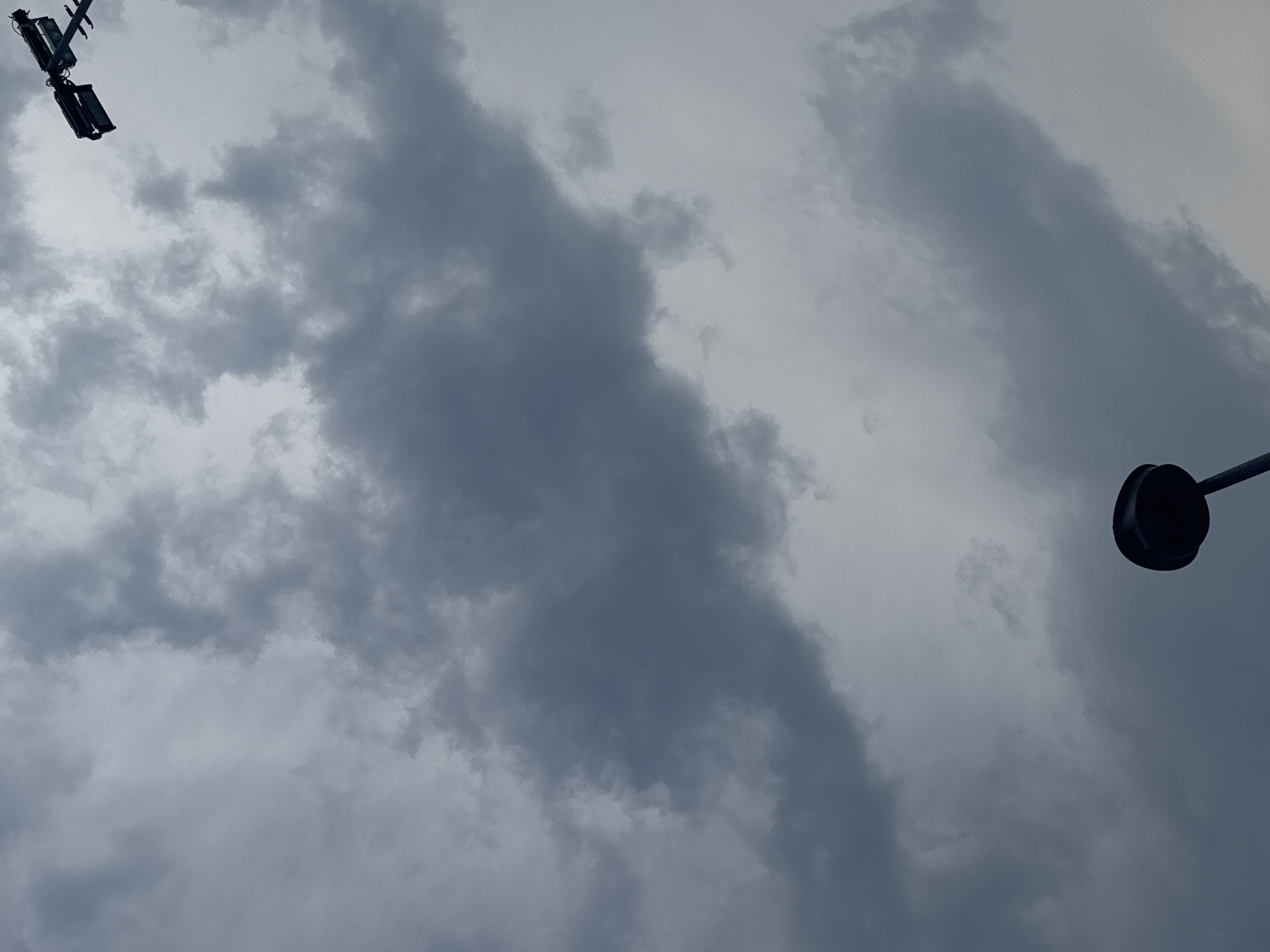The image shows a cloudy sky with two streetlights visible. One streetlight is in the upper right corner and appears to be a double floodlight. The other streetlight is in the lower center and has a circular dome shape. The sky is overcast with dark gray clouds dominating the frame, suggesting a potentially stormy or overcast day. The scene appears to be outdoors, with the sky as the primary focus. No people or specific landmarks of Chiasso, Switzerland are discernible in the image.