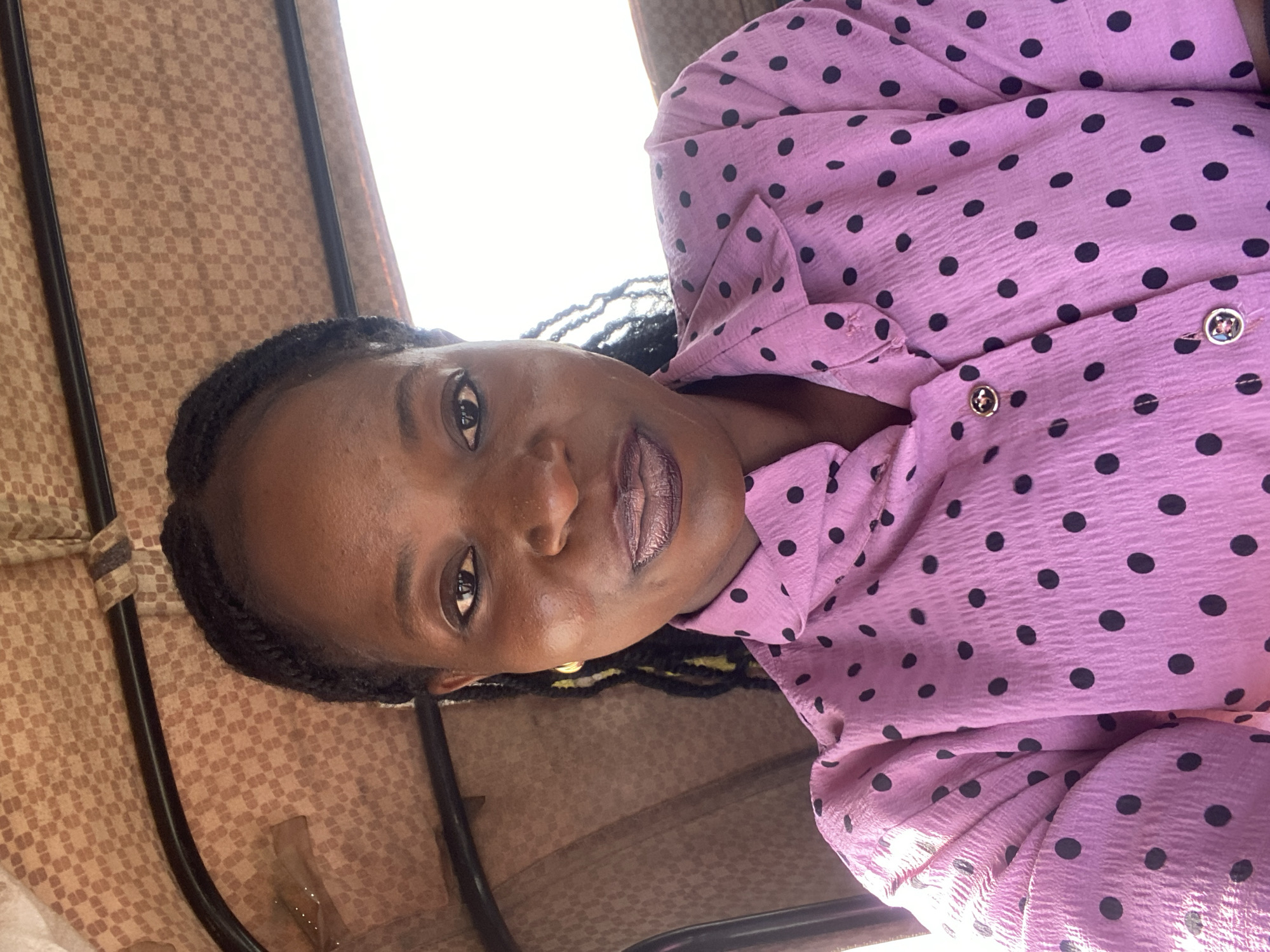 A medium shot captures a Black woman with braided hair and dark makeup, wearing a pink polka dot shirt, inside a vehicle. The woman is positioned centrally in the frame, with her face turned slightly towards the camera. Her expression is neutral. The interior of the vehicle, likely a tricycle or tuk-tuk, is visible behind her, featuring a checkered brown fabric lining. The background is largely out of focus, with a bright white area suggesting an open window or sunlight. The image is taken from a slightly upward angle. The location is identified as Bukuru, Nigeria.