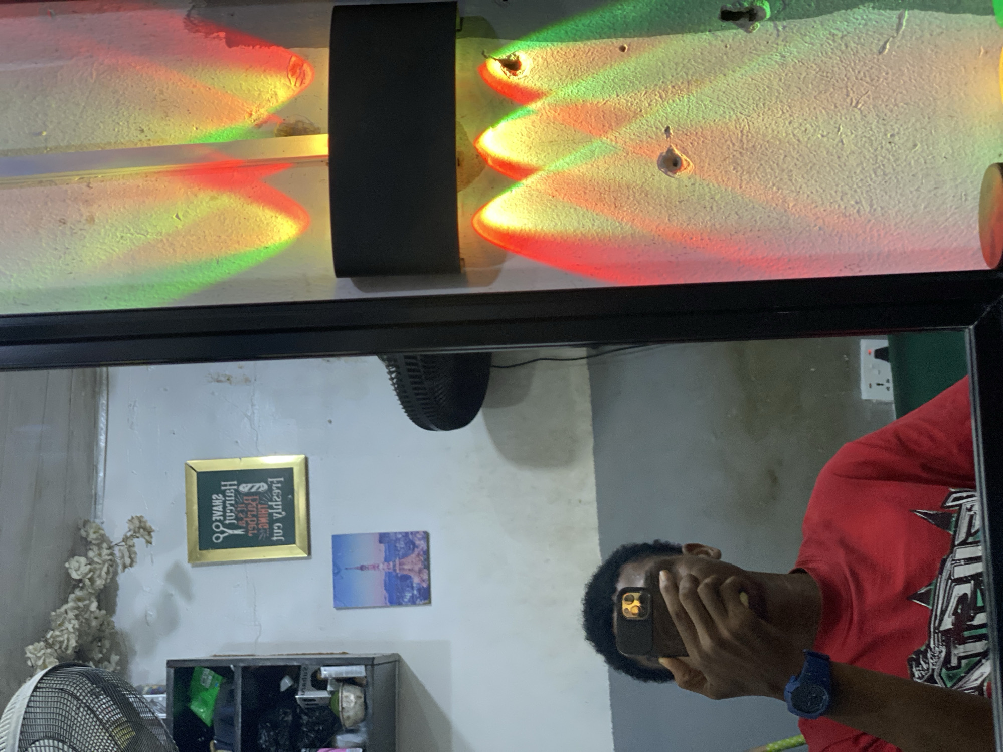 The image is a mirror selfie taken in a room. A Black man wearing a red T-shirt and a blue wristwatch holds a smartphone up to his face, obscuring his features. The room contains a fan, a small black cabinet, a framed picture with text, and a picture of a blue sky. There is a black object and decorative lighting with red and green colors above. There is no specific information confirming this is in Ado, Nigeria.