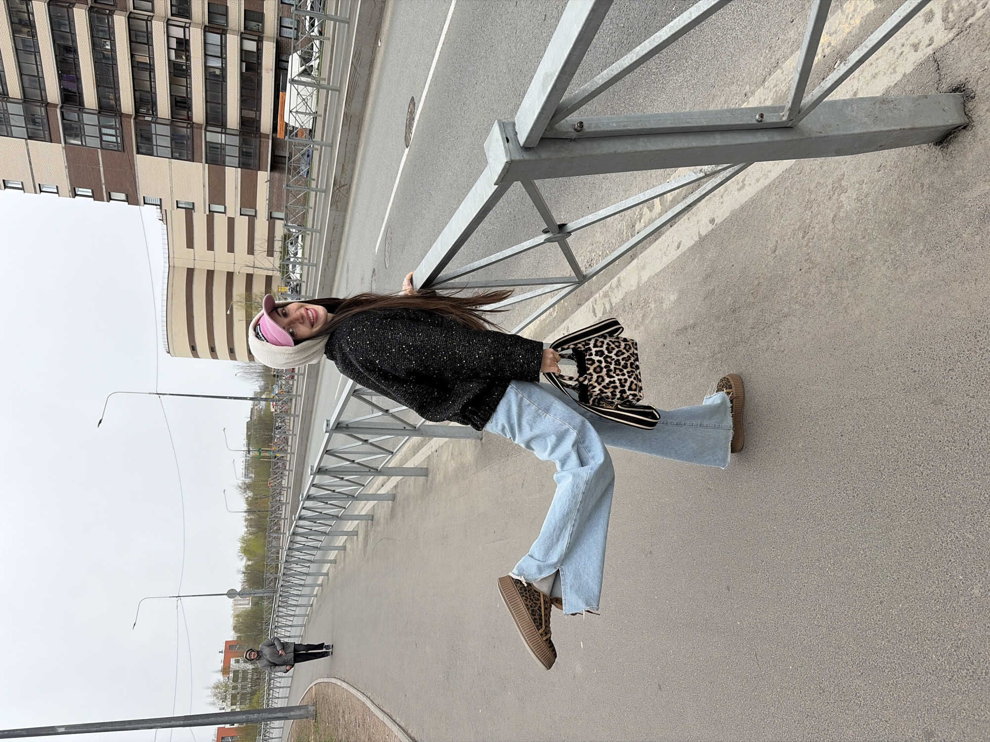 A woman in a black, speckled sweater, light blue jeans, and leopard-print platform sneakers poses on a pedestrian bridge in Saint Petersburg, Russia. She is holding onto a gray metal railing that borders the bridge, carrying a leopard-print handbag. She wears a white and pink hat. In the background is a large apartment building with a beige and brown striped facade. A street with white lines runs parallel to the bridge. The sky is overcast. A figure is walking in the distance.