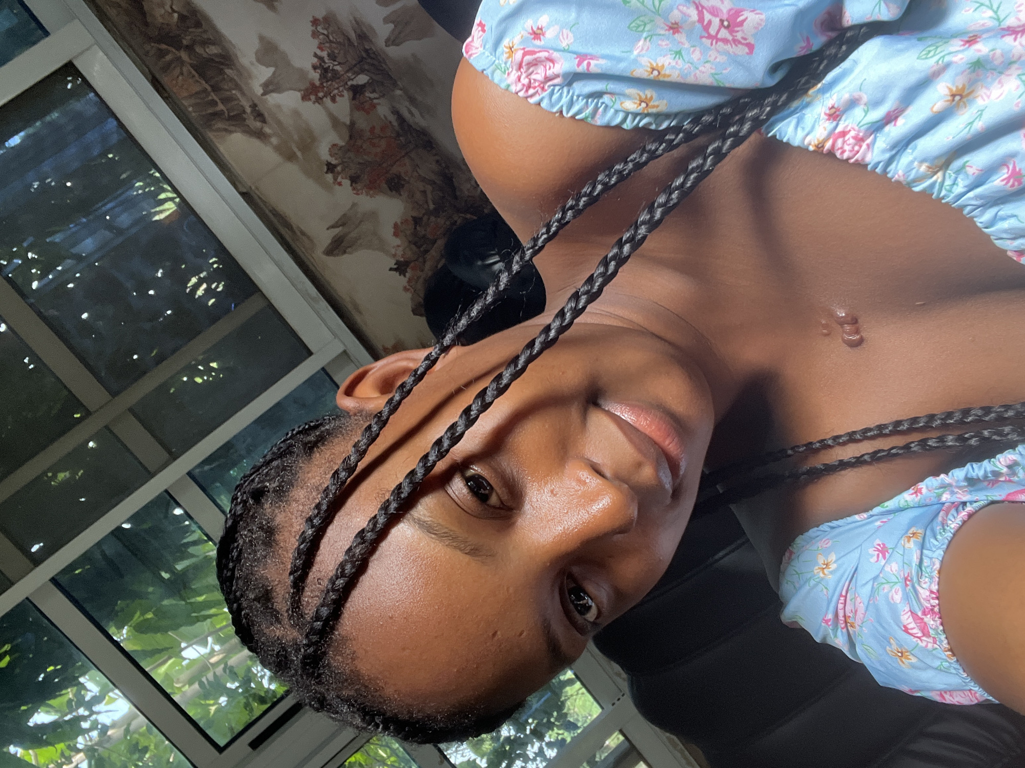 A close-up, rotated perspective photograph shows a dark-skinned person wearing a light blue floral patterned top with braids, positioned against a backdrop of a window. The person is positioned in front of the window. Reflected light from the window is visible on the subject's face and surrounding area. Part of a wall with floral-patterned wallpaper is visible. The location is presumed to be in Adankolo, Nigeria.