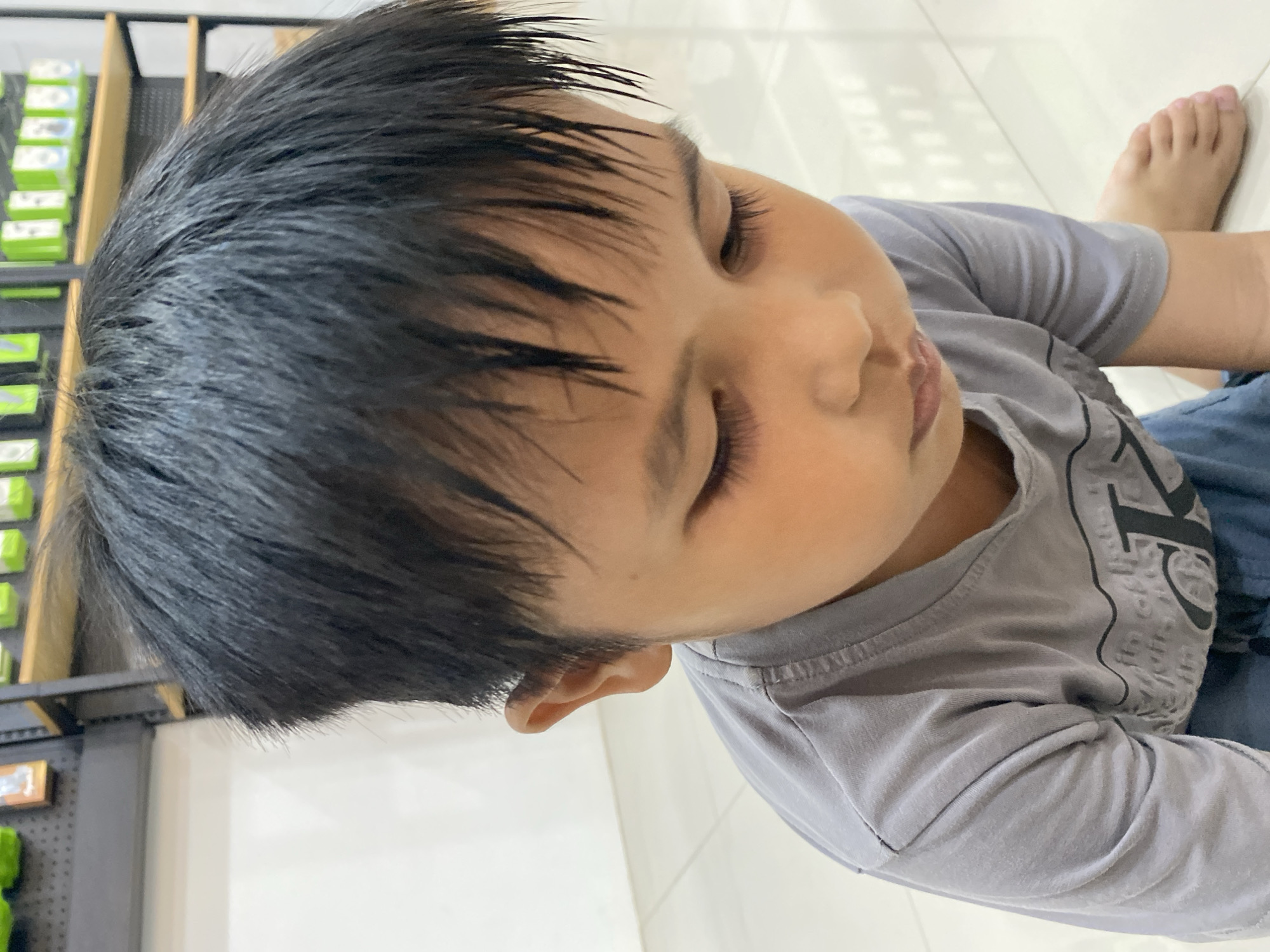 A close-up, eye-level shot shows a young boy of Southeast Asian descent with dark hair, looking down with his eyes partially closed. He is wearing a plain gray t-shirt and blue shorts. The boy is positioned on a light-colored, reflective floor, possibly tiled, in an indoor setting. In the background, out of focus, are shelves displaying numerous items, some of which appear to be packaged goods in green and white. The lighting suggests a commercial or retail environment, possibly a shop or a display area. The image does not provide specific clues about the location within Ta', Indonesia, beyond it being an indoor commercial space.