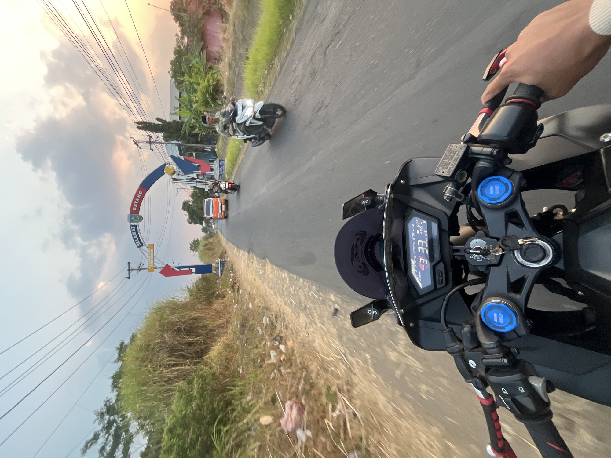 A first-person perspective shot captures a motorcycle ride towards an archway marking the entrance to Karangrandu, Indonesia. The road is asphalt with a dirt shoulder lined with dry vegetation and scattered litter. Overhead, power lines crisscross the sky, which is filled with scattered clouds, reflecting the warm glow of the setting sun.
The motorcycle's dashboard displays digital readings, including "33" in large numbers, and "5:25" and "4:45" in smaller text. The handlebars are black with red accents and a hand grips the right handlebar. In the distance, a silver scooter carrying a man in a patterned shirt and head covering rides towards the camera. To the left, an orange truck and another motorcycle are visible on the road. Beyond the road, buildings with pink and white exteriors and lush green trees are present. The archway itself features blue and red elements with Indonesian text that reads "SELAMAT DATANG," meaning "Welcome."
