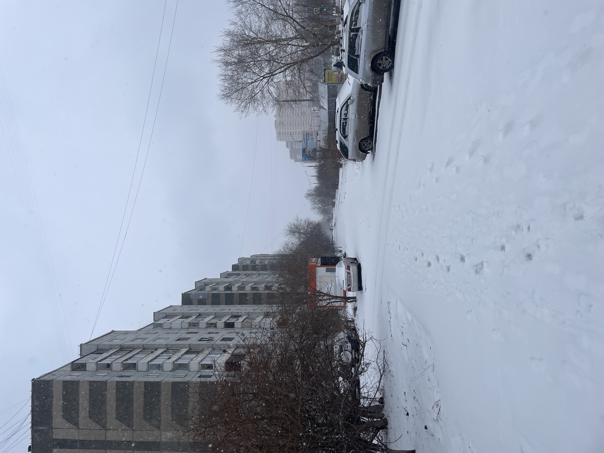 The scene is a snowy street in Krasnoyarsk, Russia. A tall, grey apartment building with a stepped design is on the left side of the frame. On the right side of the street, two light-colored cars are parked alongside a snow-covered curb. In the center of the frame, a white van with an orange stripe is parked further down the street. The road and sidewalks are covered in snow with visible footprints. Bare trees and bushes line the street between the buildings and cars. Power lines stretch across the sky. In the distance, more apartment buildings are visible through the haze. The sky is overcast and snow is falling.