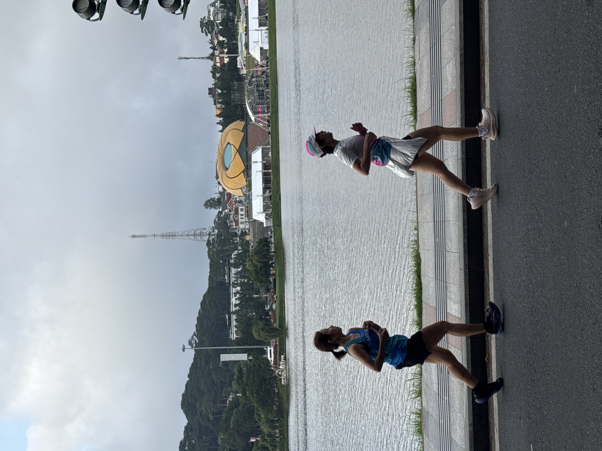 Two adult females are jogging on an asphalt road alongside a body of water in Phường Xuân Hương, Đà Lạt, Vietnam, under an overcast sky.
The scene is dominated by a large body of water, likely Xuân Hương Lake, which occupies the midground. A narrow strip of green grass separates the water from a pedestrian sidewalk. The sidewalk, composed of grey rectangular pavers, includes sections with tactile paving (white and pinkish-brown ridged slabs). The asphalt road occupies the bottom portion of the image, where the two individuals are jogging from left to right.
The woman on the left has light brown hair tied in a ponytail and wears a blue and white patterned sleeveless athletic top with the partial text "UPR" visible on the chest, black athletic shorts, and dark-colored running shoes with light-colored soles. She holds a small, clear bottle in her right hand.
The woman on the right wears a white short-sleeved top, a multi-colored (pink, blue, white) fanny pack or waist bag, light-colored shorts or a skirt-like overlay, black athletic shorts beneath, and light pink running shoes with white soles. She wears a multi-colored (pink, blue, white) baseball cap.
In the background, behind the lake, is a panorama of densely forested hills. On the left, several multi-story buildings, including a prominent white structure, are visible among the trees. A tall, grey street light pole stands near the trees. Further right, a distinctive large, circular, golden-colored building with a blue central feature, resembling a lotus, stands near the water's edge. This structure is consistent with Lâm Viên Square. Adjacent to this building and closer to the lake, several white temporary event tents or structures are set up. Two slender transmission towers, one red and white, the other grey, are visible on distant hills. A traffic light pole with three dark, round light fixtures is partially visible on the far right edge of the image. The sky is uniformly cloudy and grey.
