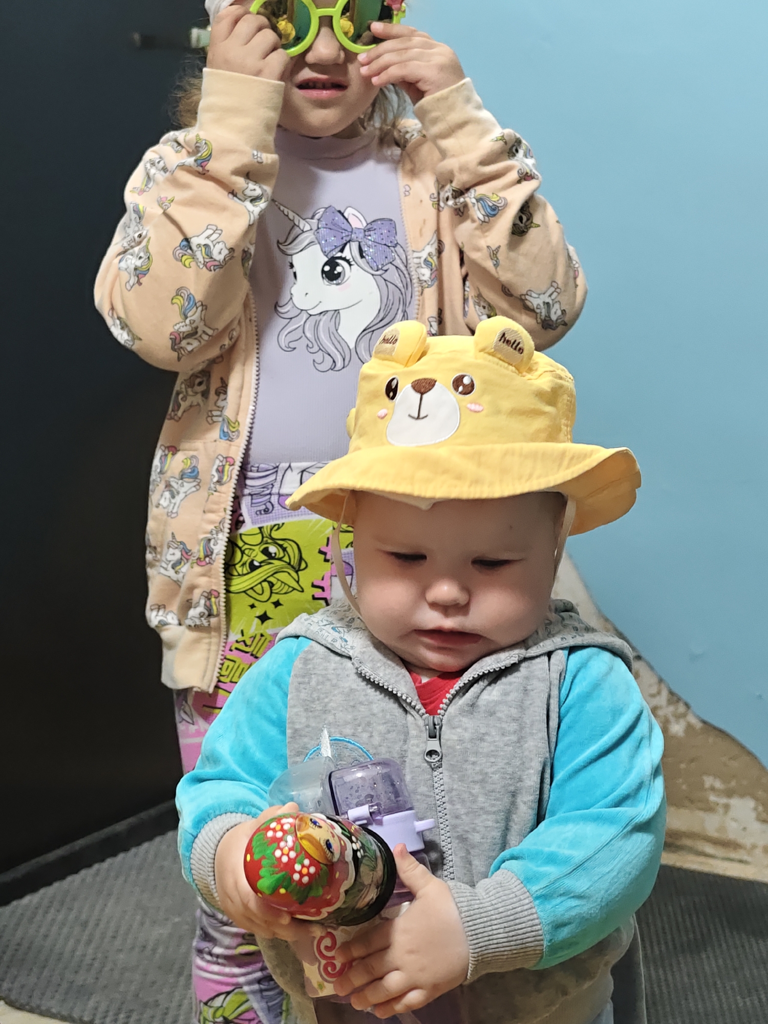 Two children are present. In the foreground, a baby wears a yellow hat with a bear face and is holding a set of Russian nesting dolls. The baby is wearing a zip-up jacket in teal and gray. In the background, an older child stands wearing sunglasses and a jacket with unicorn images on it, over a shirt with a unicorn. They are in front of a blue wall. The photograph appears to be taken in an indoor location. The exact location in Chaikovsky, Russia is unclear.