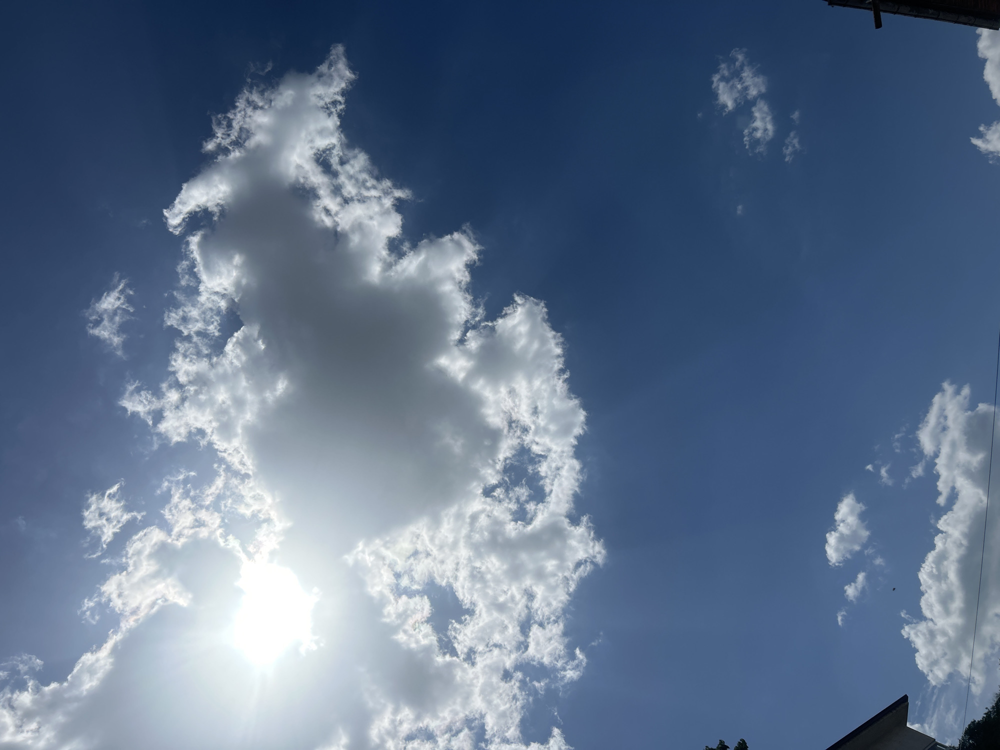 The image shows a daytime sky in Unknown, Unknown, featuring a blue background with varying shades due to sunlight and atmospheric perspective. There are several white, fluffy cumulus clouds, with a large one dominating the left side of the frame. The sun is behind this cloud, causing a bright glare and radial sunbeams. Smaller clouds are scattered across the right side of the image. The very edges of buildings are visible at the bottom of the shot.