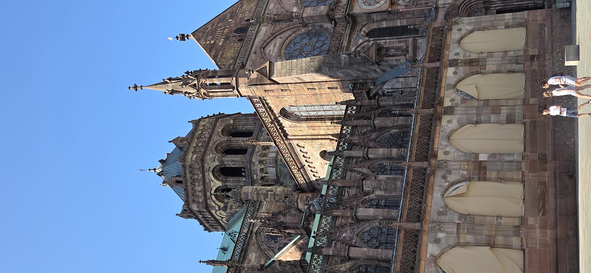 The image shows a low-angle, vertical shot of the Strasbourg Cathedral, officially known as the Cathédrale Notre-Dame de Strasbourg, in Strasbourg, France. The cathedral's Gothic architecture is prominent, with its reddish-brown sandstone facade, intricate carvings, and stained-glass windows. The sky above is a clear, bright blue.
In the foreground, a group of three young people, two females and one male, are visible. They are casually dressed in t-shirts and shorts. The male is on the left, wearing a white t-shirt and dark blue shorts. The two females are to his right, both wearing white t-shirts and light-colored shorts. They appear to be interacting, possibly playing or walking. Small white particles are scattered in the air around them, suggesting they might be playing with water or some other small objects.
The cathedral occupies the majority of the frame, showcasing its imposing scale and detailed construction. The lower part of the facade features large arched windows, some of which are covered with beige tarps, possibly for restoration or protection. Intricate stone tracery and decorative elements are visible above these covered windows. Higher up, there are more arched windows, including rose windows, and ornate spires topped with crosses. The sunlight casts shadows on the building, highlighting its architectural features. The ground in front of the cathedral appears to be paved.