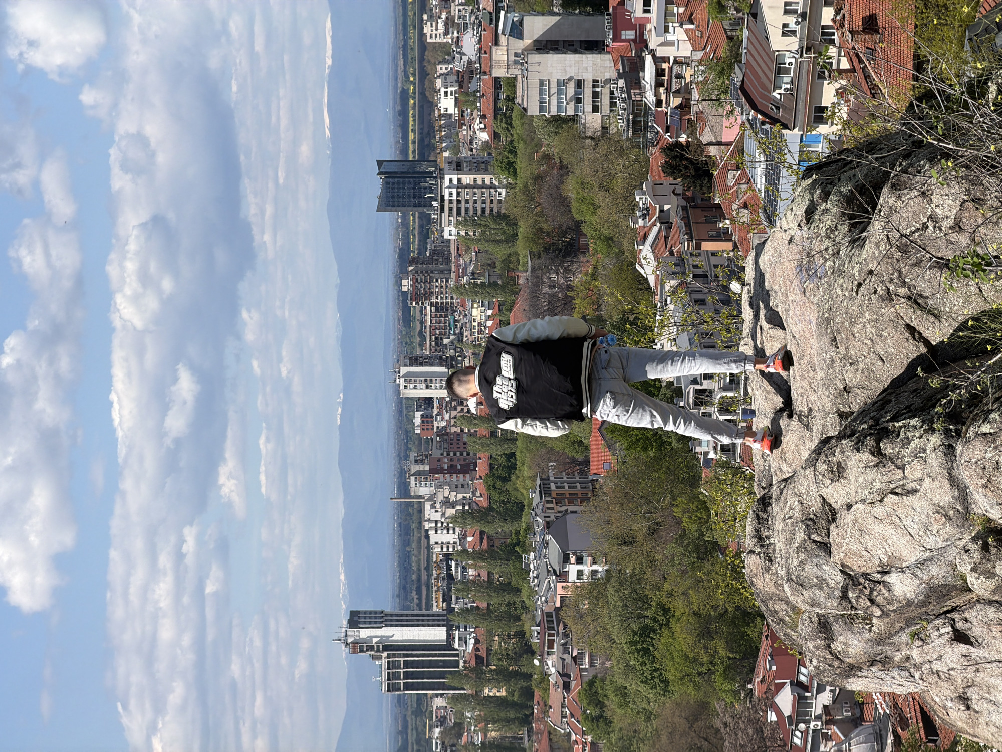 A vertically-oriented photograph shows a person standing on a rock overlooking the city of Plovdiv, Bulgaria. The sky is blue with patchy clouds and the skyline is dotted with mountains. The person is wearing gray pants and a black jacket with white sleeves. The rock is textured and gray, and the background shows a densely packed city with buildings of varying heights and designs. There are trees interspersed among the buildings.