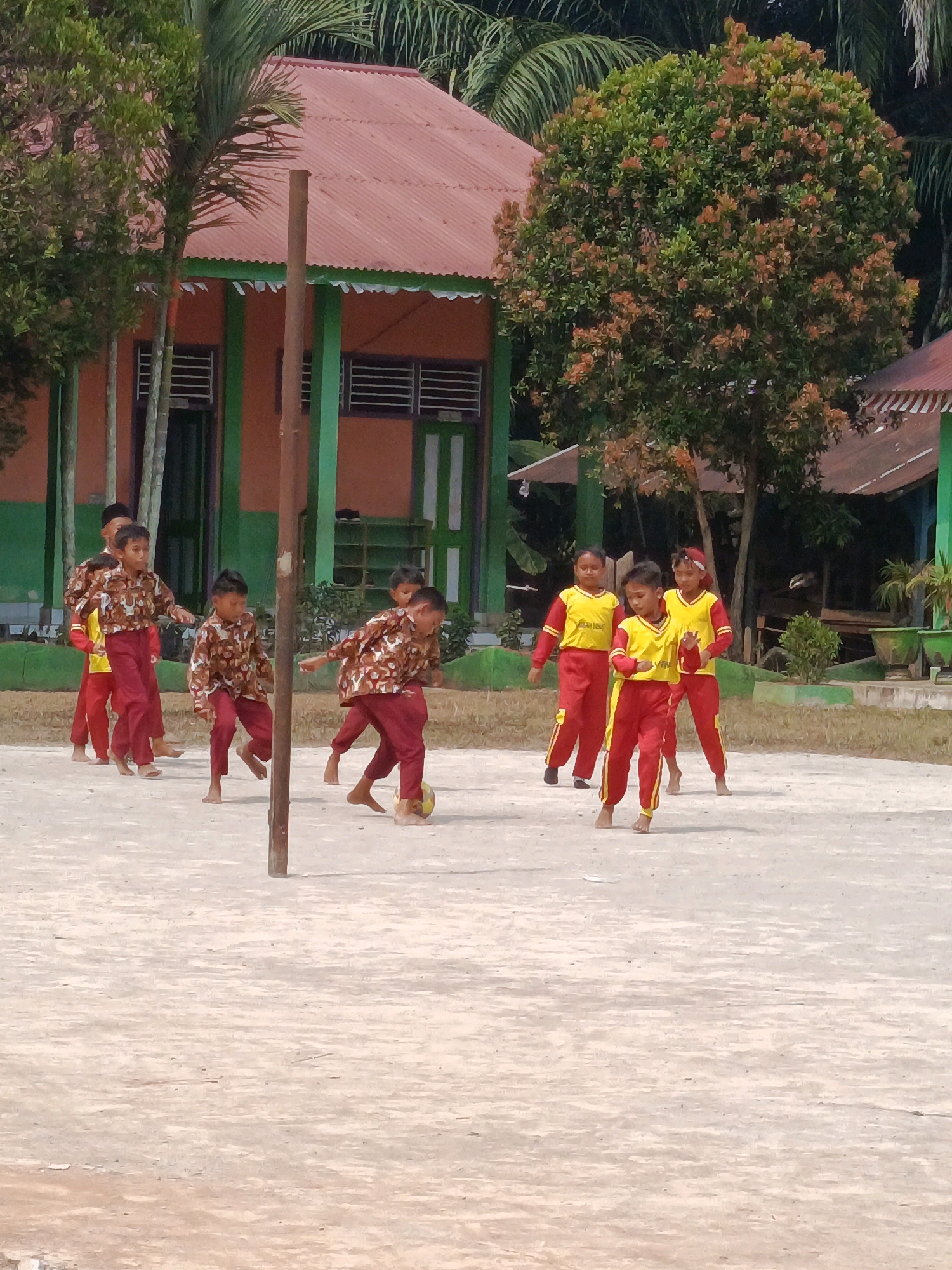 This is a medium-angle, eye-level, full shot of a group of children playing soccer barefoot on a sandy playground in front of a low-rise building, likely a school, with a red corrugated metal roof and green trim. The building has several windows with louvers and open doorways. Lush green trees with some reddish-orange foliage are visible behind and to the side of the building. The playground surface is a light brown, sandy dirt.
Several children are actively engaged in playing soccer. Two children in the foreground are wearing matching patterned brown and white shirts and maroon pants, with one child kicking a yellow soccer ball. Another child to the left is wearing a yellow shirt and red pants. Two children in the middle ground are wearing identical yellow shirts with red stripes and red pants. Further to the left, two more children are visible, one in a yellow shirt and red pants and another in a patterned brown and white shirt and maroon pants. The children's expressions and body language suggest active play and exertion. The presence of a single wooden pole in the foreground indicates the area may be used for sports or other activities. The overall scene depicts a lively outdoor activity, likely during daylight hours, given the illumination. The setting appears to be a rural or semi-rural area, consistent with a location like Megang Sakti IV, Indonesia.