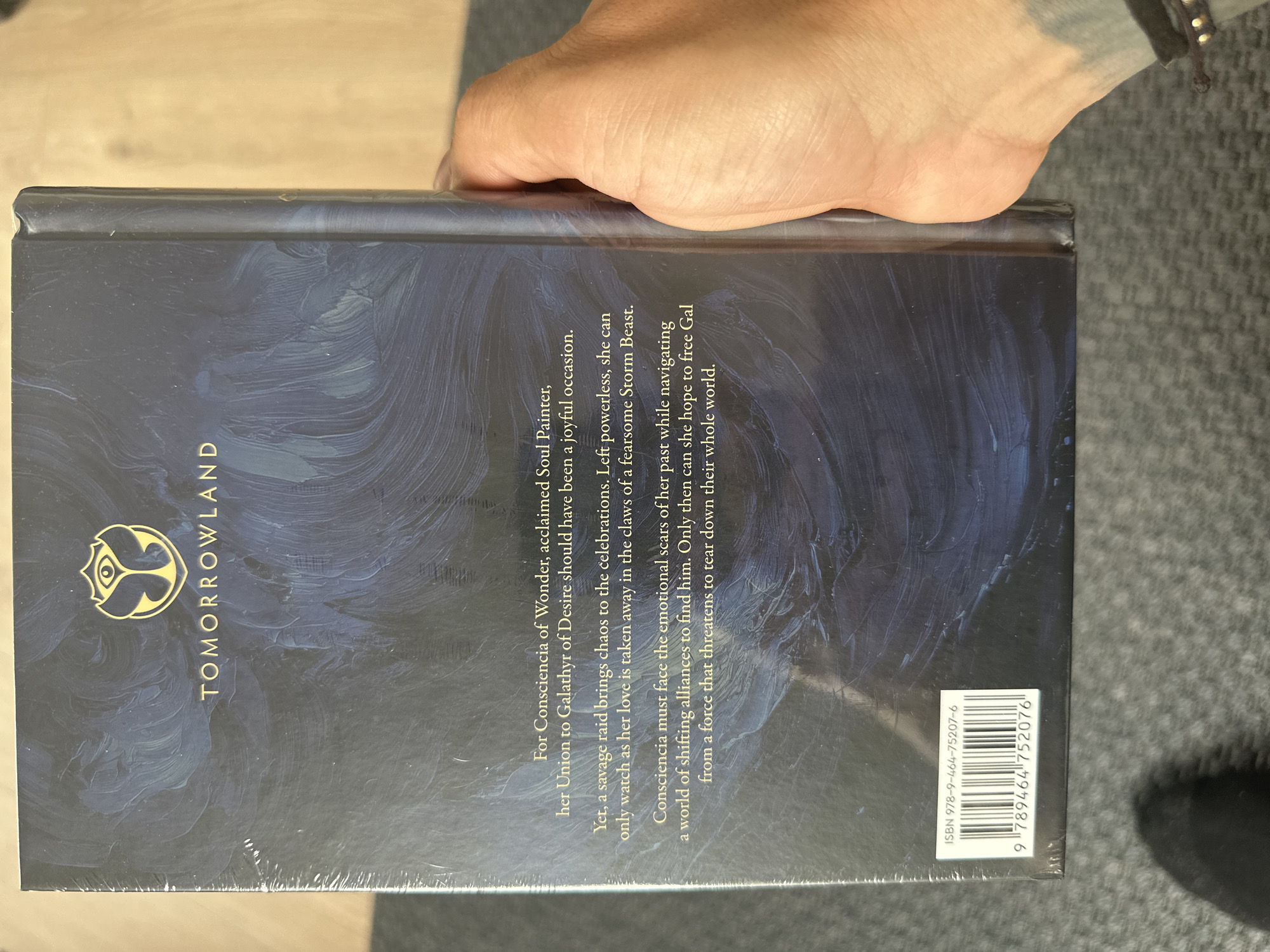The image shows the back cover of a book titled "Tomorrowland." The cover is dark blue with swirling patterns resembling brushstrokes. In the upper left corner is a golden logo, and next to it is the title "TOMORROWLAND" in vertical lettering. The back cover contains a blurb about the story. The ISBN is 978-9-464-75207-6. The barcode is 9789464 752076. A human hand is partially visible at the top of the frame. The surface beneath the book is gray. The city of Nivelles and Belgium is not detectable in the image.