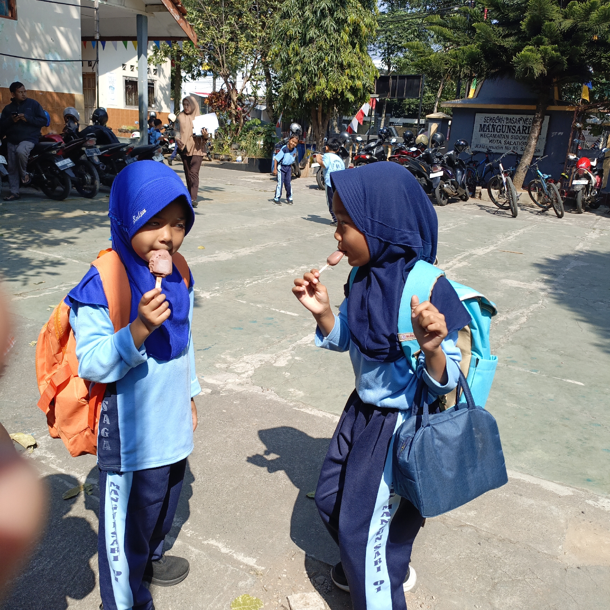 Two young girls in blue hijabs and light blue school uniforms are standing on a paved outdoor area, likely a schoolyard in Salatiga, Indonesia. The girl on the left, wearing an orange backpack, is eating a chocolate popsicle. The girl on the right, with a turquoise backpack, is also eating a popsicle, sticking her tongue out. The background shows parked motorcycles, bicycles, trees, and parts of a building with signage that reads "SEKOLAH DASAR NEGERI MANGUNSARI" and "KECAMATAN SIDOMULYO KOTA SALATIGA". The lighting suggests it is a sunny day.