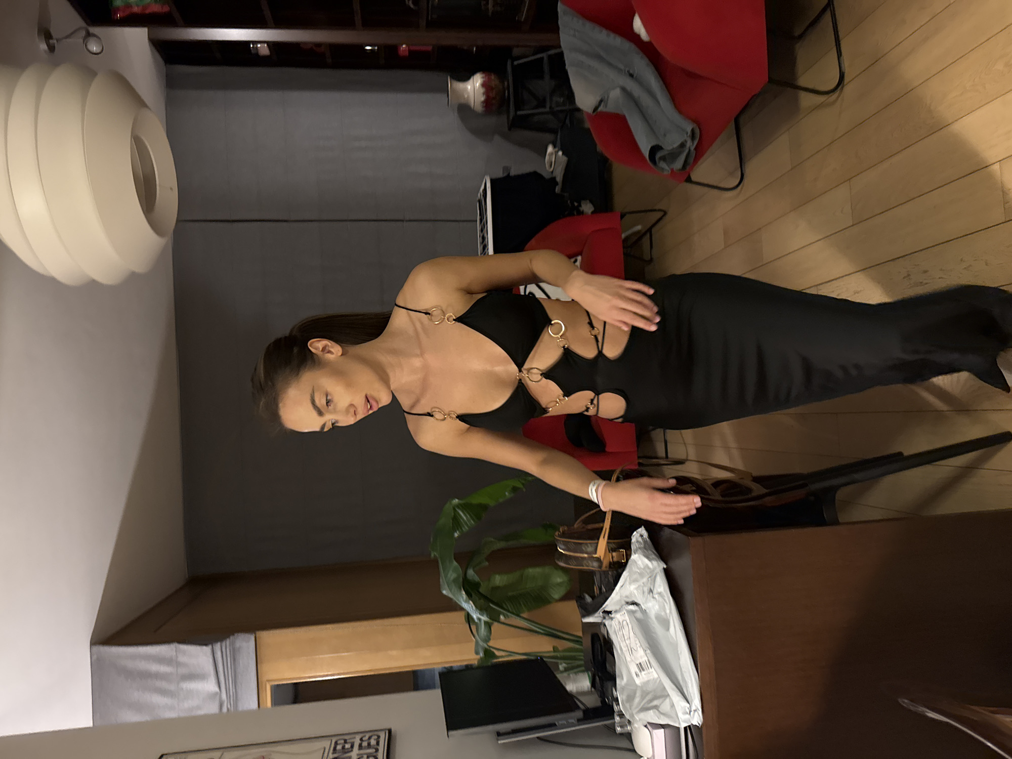 A woman is standing in an interior. She is wearing a black dress with cutouts and gold ring detailing. Her hair is pulled back in a ponytail. She is reaching towards a table with a small brown handbag and a plant on it. A monitor, packaging, and other items are on the table as well. There is a red chair and other furniture in the background, including a large modern white lamp. The location is most likely a private residence in Warsaw, Poland.