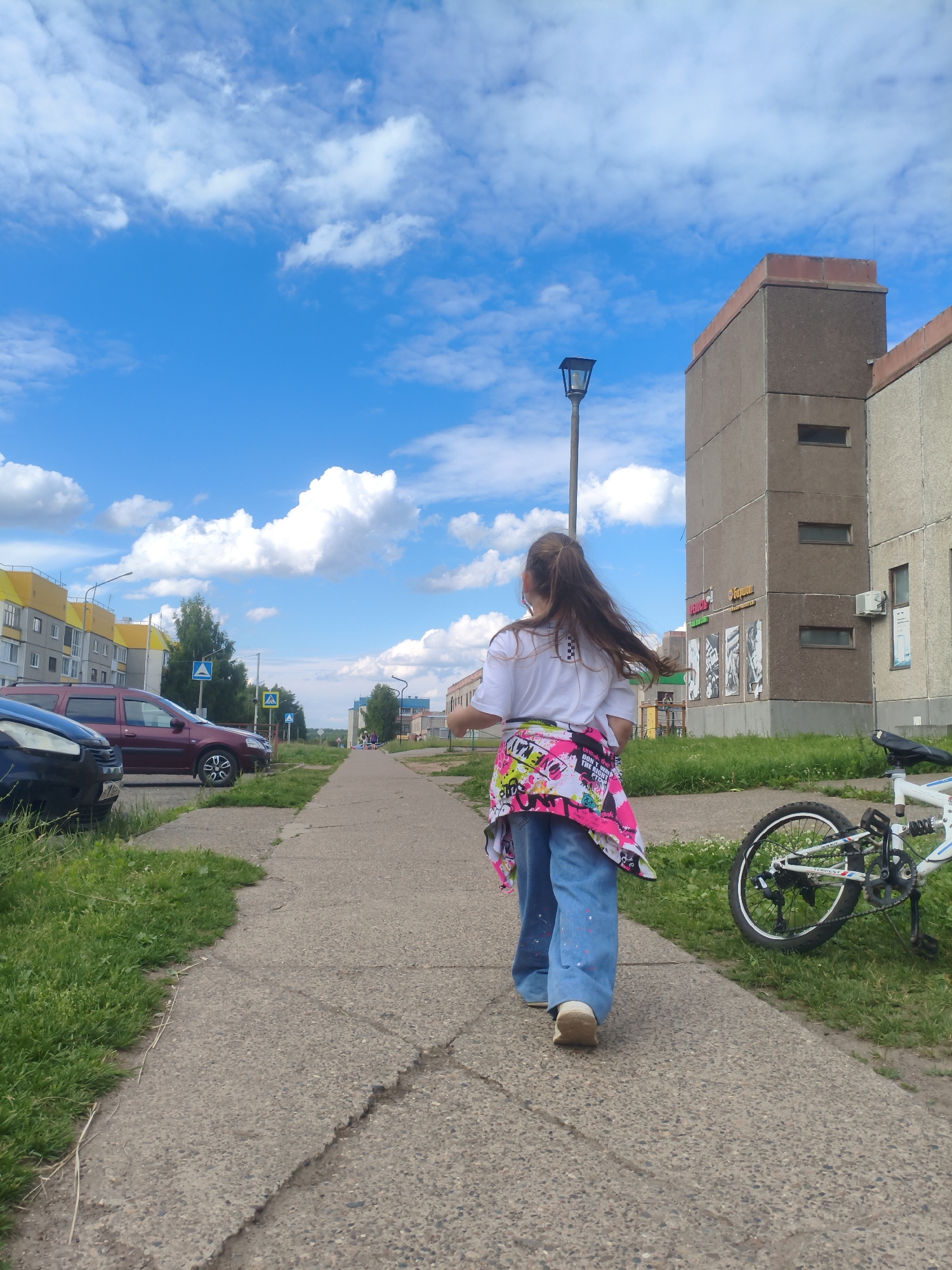 A young girl with long brown hair is walking away from the camera down a concrete sidewalk. She is wearing a white t-shirt, loose-fitting blue jeans, and beige shoes. A brightly colored jacket with a graffiti-like pattern is tied around her waist. Beside the sidewalk, to the right of the girl, a white bicycle is lying on its side on the grass. The scene takes place in an urban environment with multi-story buildings visible in the background. A street lamp stands on the left side of the sidewalk. Cars are parked along the left side of the street, and grass and trees line the sidewalk. The sky is bright blue with scattered white clouds. The location appears to be in Марковский, Russia, as indicated by the presence of apartment buildings and cars typical of the region.