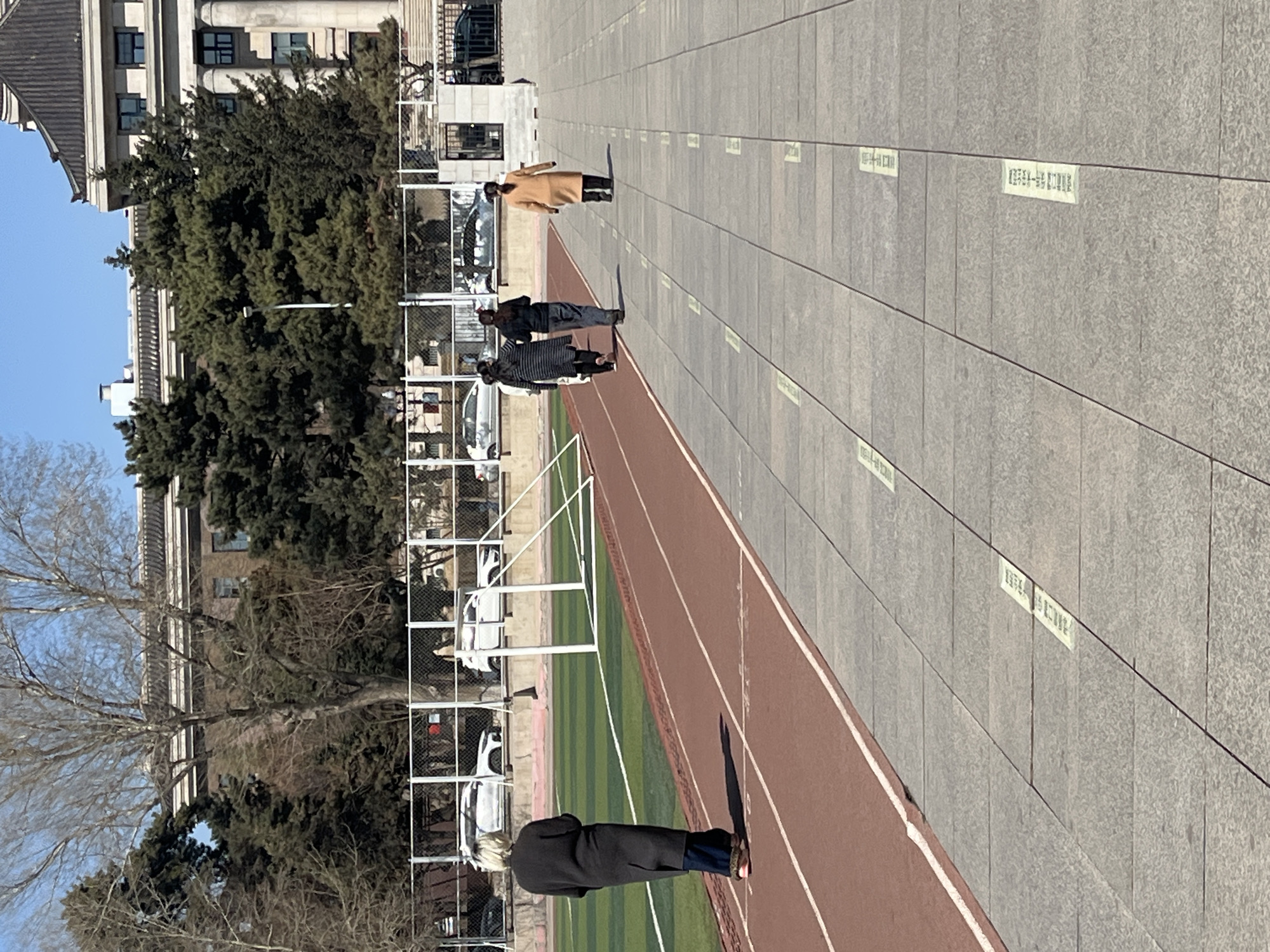 The image depicts a group of people walking along a tiled walkway adjacent to a running track. The walkway has rectangular tiles, and some have white labels affixed. The running track is red with white lines and is next to a green artificial turf field enclosed by a white fence. A building with a classical architectural style is visible in the background behind bare trees and evergreen trees. Parked vehicles are also seen behind the fence. Based on the style of architecture and appearance, the location is likely within the campus of Jilin University, located in Changchun City, China.