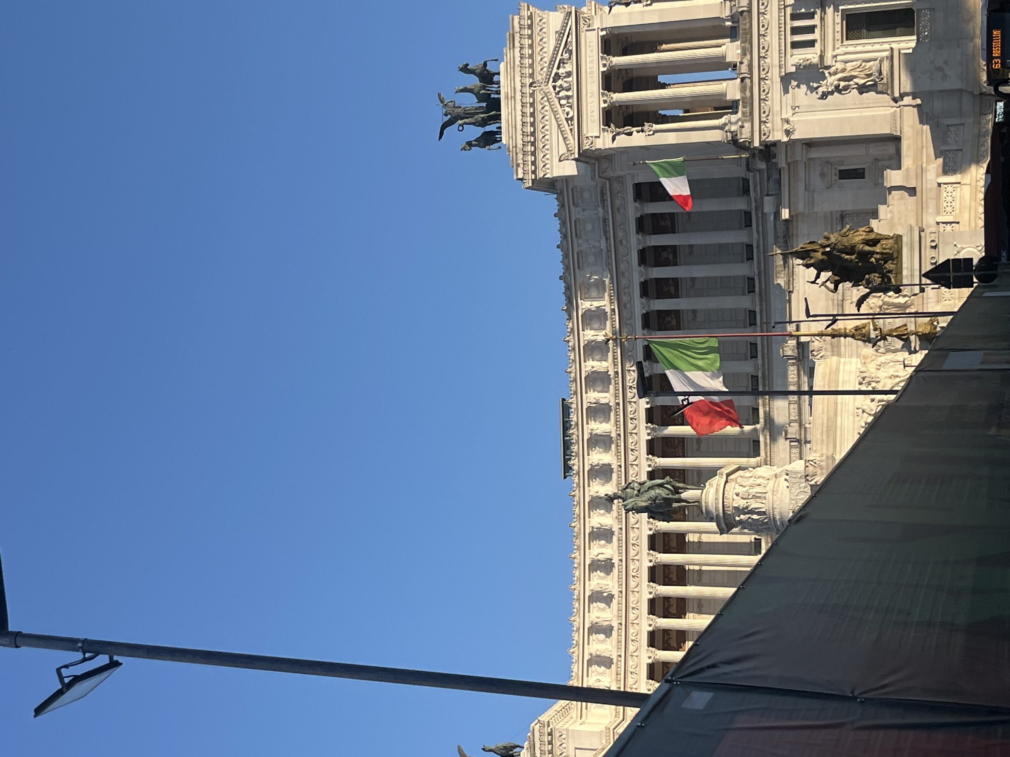 The image displays a clear, sunlit view of a significant portion of the Victor Emmanuel II National Monument, also known as the Altare della Patria, located in Rome, Italy. The monumental building, constructed from white marble, stands grandly against a vibrant, cloudless blue sky, indicating fair weather during the daytime.
Architectural details are prominent, including numerous classical columns, intricate carvings, and decorative friezes. Several Italian flags, characterized by their green, white, and red vertical stripes, are seen flying from various points on the building's facade. On the upper tier, large bronze quadrigae (chariots drawn by four horses) statues are visible, along with the Latin inscription "CIVIUM LIBERTATI" (To the Liberty of Citizens) carved into the stone. Further down, another large bronze equestrian statue on a pedestal and various sculptural reliefs adorn the monument.
In the upper right corner, partially visible, is a sign that reads "63 ROSSELLINI," possibly indicating a street name or bus route. The lower right portion of the monument is partially obscured by a dark, taut fabric or tarp structure, which might be part of scaffolding, an event setup, or an advertisement. A metal pole with an attached light fixture extends into the sky from the lower left foreground. No people are distinctly visible in the frame, and no specific event or activity is taking place beyond the static presentation of the monument.