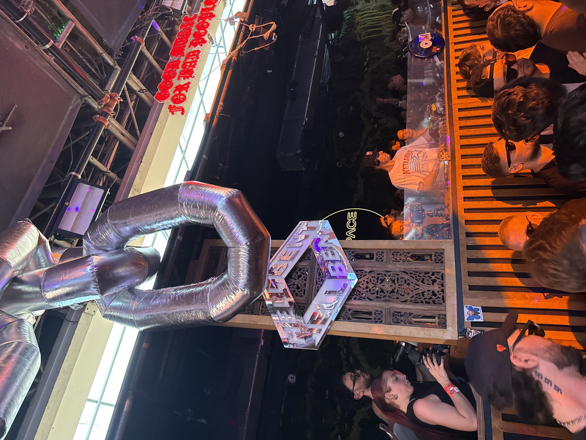 The image depicts a crowded indoor nightclub scene at Club Space in Miami, Florida, United States of America.
Overhead, large, silver-colored, reflective soft-sculpture chain links are suspended from an exposed ceiling, which features metal beams, pipes with red fittings, electrical conduits, and fluorescent strip lighting. A rectangular stage light with a grille is visible. A red neon sign reads "GOOD MUSIC FOR BAD PEOPLE," with a "SPACE" neon sign below it.
In the midground, an iridescent hexagonal sign, displaying "HELLBENT" on its faces, hangs prominently near a DJ booth. The DJ booth features a transparent protective barrier and a wooden slatted front. A male DJ wearing a white "KETAMINE USA CMHC" t-shirt, a black baseball cap, and headphones is operating Pioneer CDJ players and a mixer. Another individual stands behind the DJ. To the left of the DJ booth, a woman with reddish-brown hair, wearing a black top and a wristband, leans over a wooden railing, holding a professional camera with an attached flash unit. Behind the DJ booth, a wall covered in green foliage is visible. A blue butterfly sticker is affixed to the wooden railing.
The foreground is filled with a dense crowd of patrons. Many individuals wear sunglasses. A man in the immediate foreground, with visible neck tattoos, wears a black baseball cap and faces away from the camera. The crowd is illuminated by warm, orange-toned lighting, creating distinct highlights and shadows.