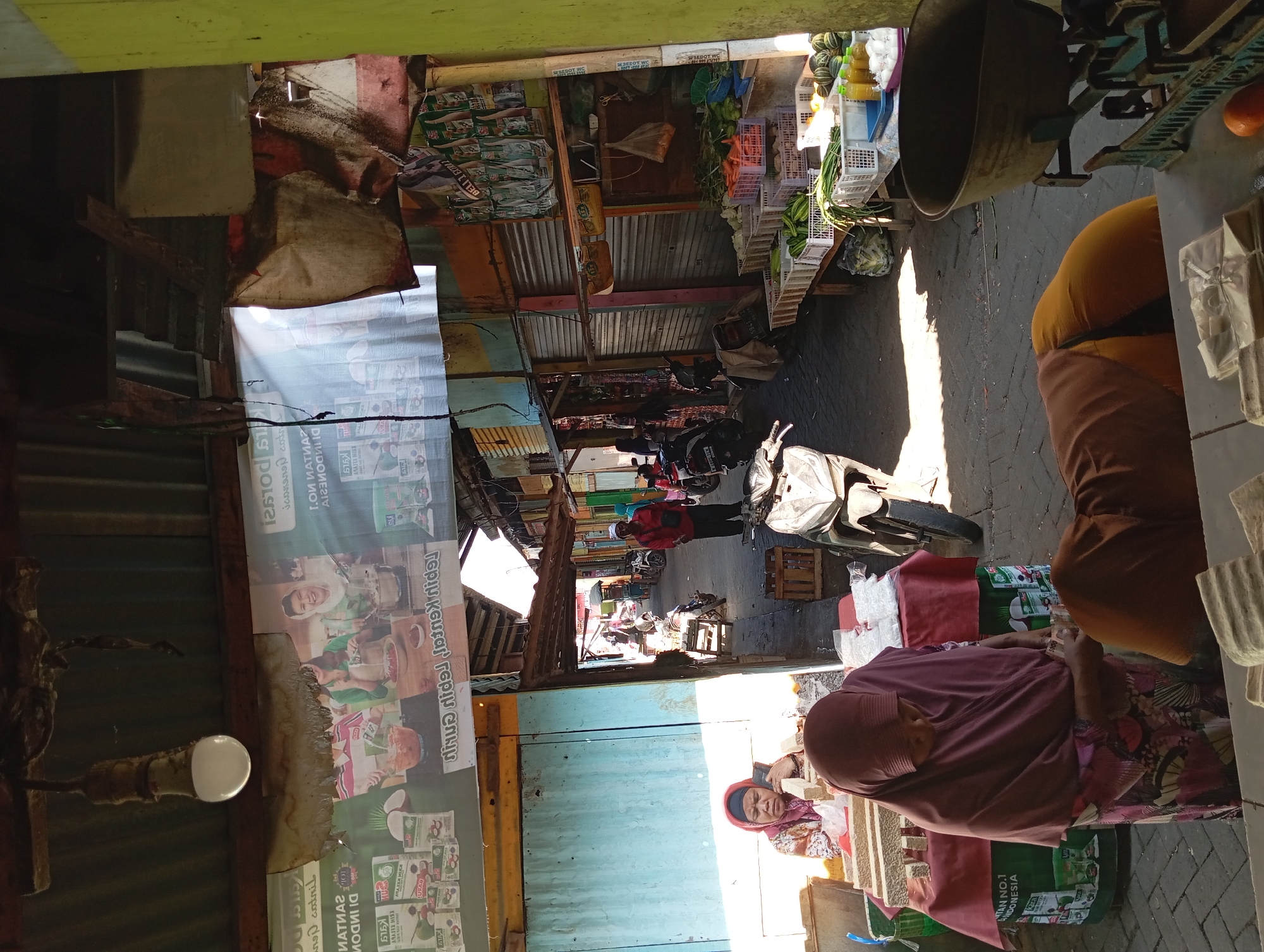 The image depicts a bustling outdoor market scene in Kepulungan, Indonesia. In the foreground, two individuals are engaged in a transaction. One, a woman wearing a purple headscarf and a patterned dress, is standing and appearing to hand over money to another person whose back is turned, wearing a brown garment and an orange undershirt. They are positioned behind a table displaying stacked, rectangular objects, possibly food items like tofu or tempeh, wrapped in white plastic. To the left, another woman, also in a headscarf and patterned dress, is seated and looking down.
The market stalls stretch into the background, characterized by corrugated metal roofing supported by wooden beams. Various goods are displayed, including baskets filled with fresh produce such as carrots, cucumbers, and melons. Bottles of liquid, likely drinks, are also visible. Several motorcycles are parked throughout the market, indicating transportation methods for vendors and customers. Advertising banners with images of food products and text are hung overhead and on the stall walls, some of which are torn and weathered. The ground is paved with cobblestones. Sunlight casts strong shadows, suggesting it is daytime. The overall environment appears somewhat rustic and active, typical of a local Indonesian market.
