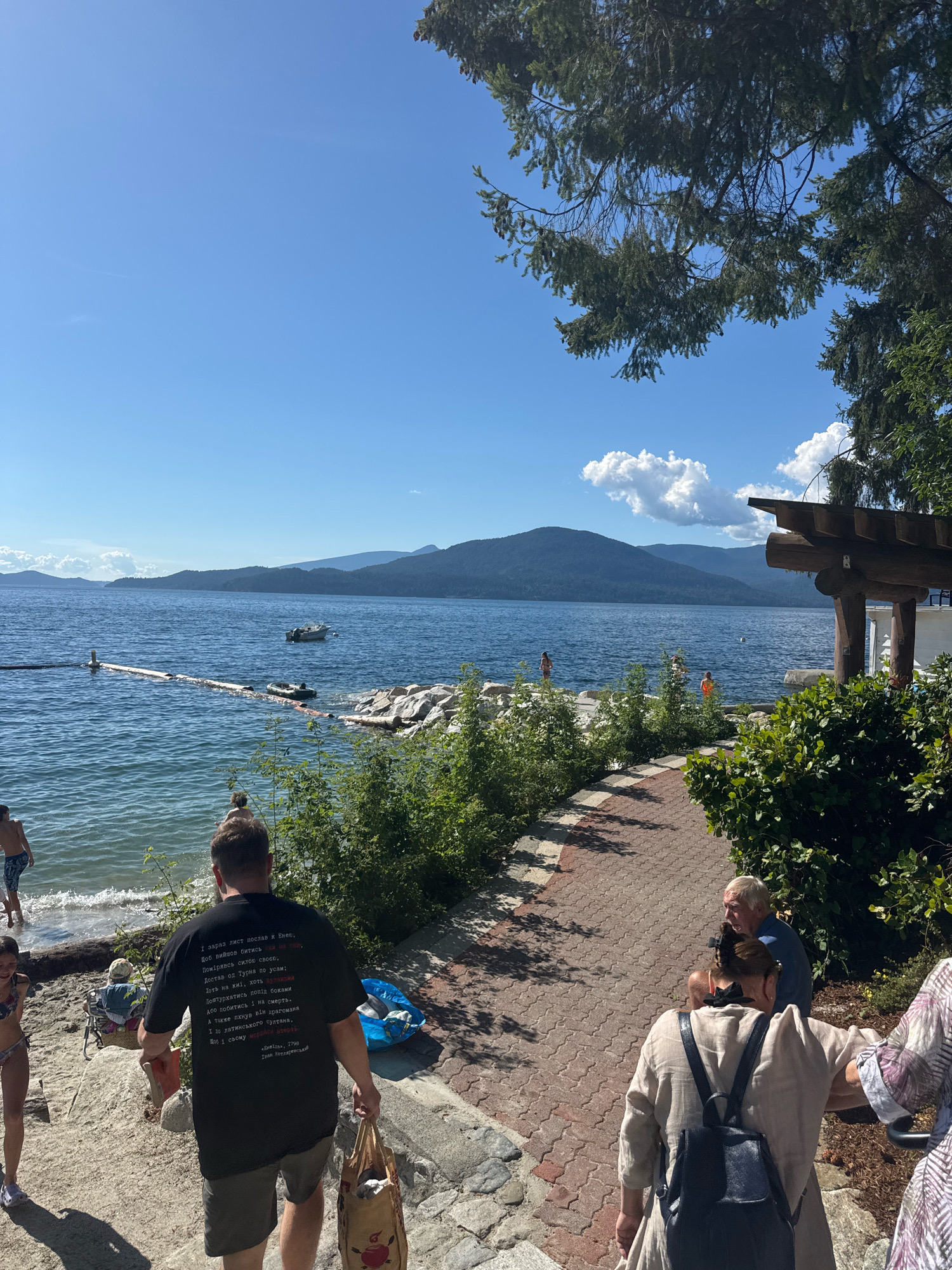 The image depicts a sunny day at a waterfront location, likely a park or beach area, in Lions Bay, Canada. In the foreground, a man in a black t-shirt with text printed on it is walking away from the camera, carrying a bag. Behind him, a woman in a bikini is visible. Further back, a person is wading in the water. To the right, a paved walkway lined with bushes leads towards a wooden structure, possibly a pavilion, where two people are seated. The middle ground shows a body of water with a boat and a floating barrier. In the background, mountains rise against a clear blue sky with scattered clouds. The overall scene conveys a leisurely, outdoor atmosphere during warm weather.