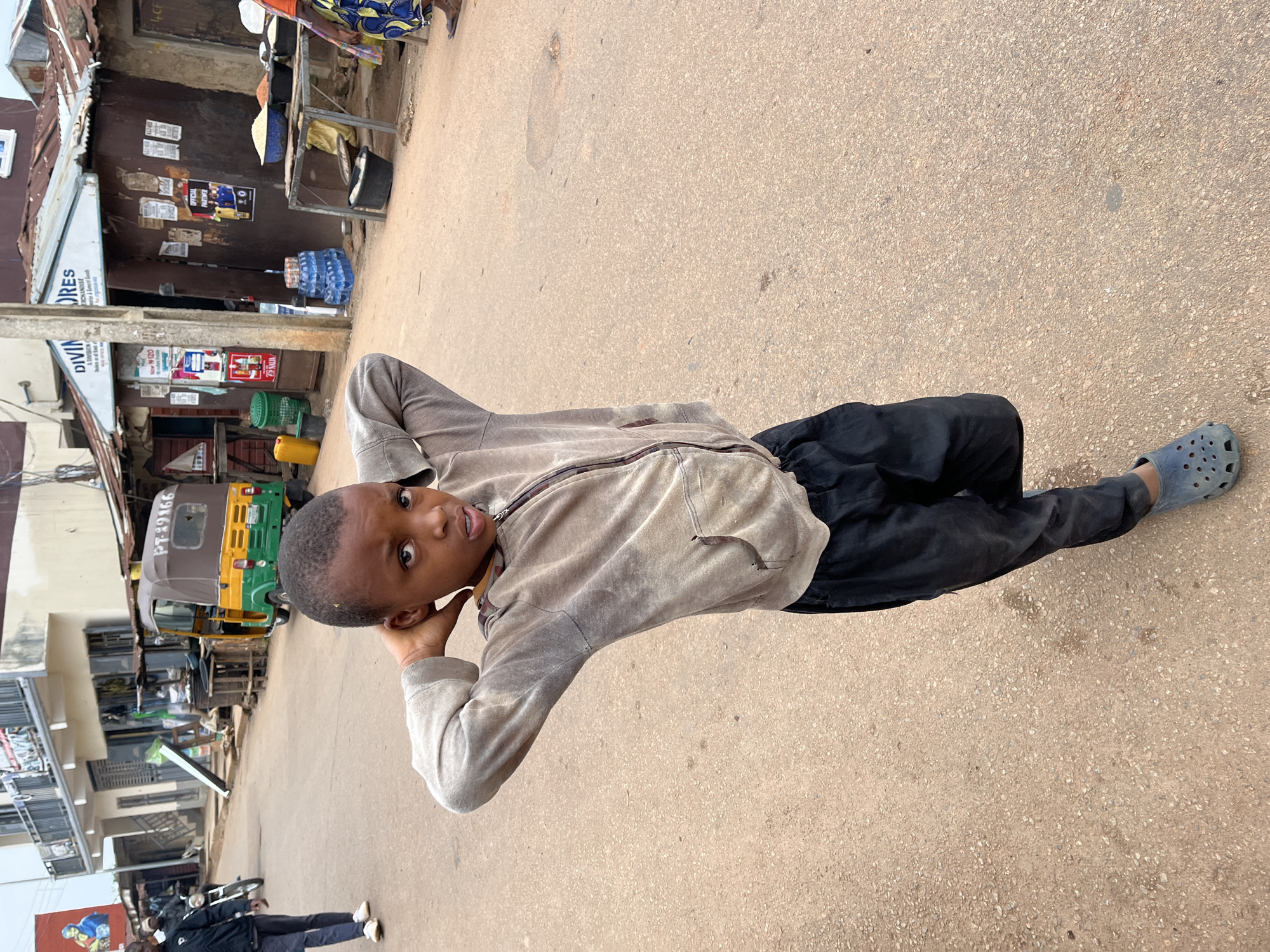 A young boy stands on a dirt road in Jos, Nigeria, with his hands behind his head. He is wearing a light brown zip-up hoodie and black pants. The boy is looking directly at the camera with his mouth slightly open. In the background, a yellow and green auto-rickshaw is parked on the side of the road. Several buildings line the street, some with corrugated metal roofs. A stall with sacks of rice is also visible, as are stacks of bottled water and other goods. The scene appears to be a busy marketplace or street in a developing urban area.