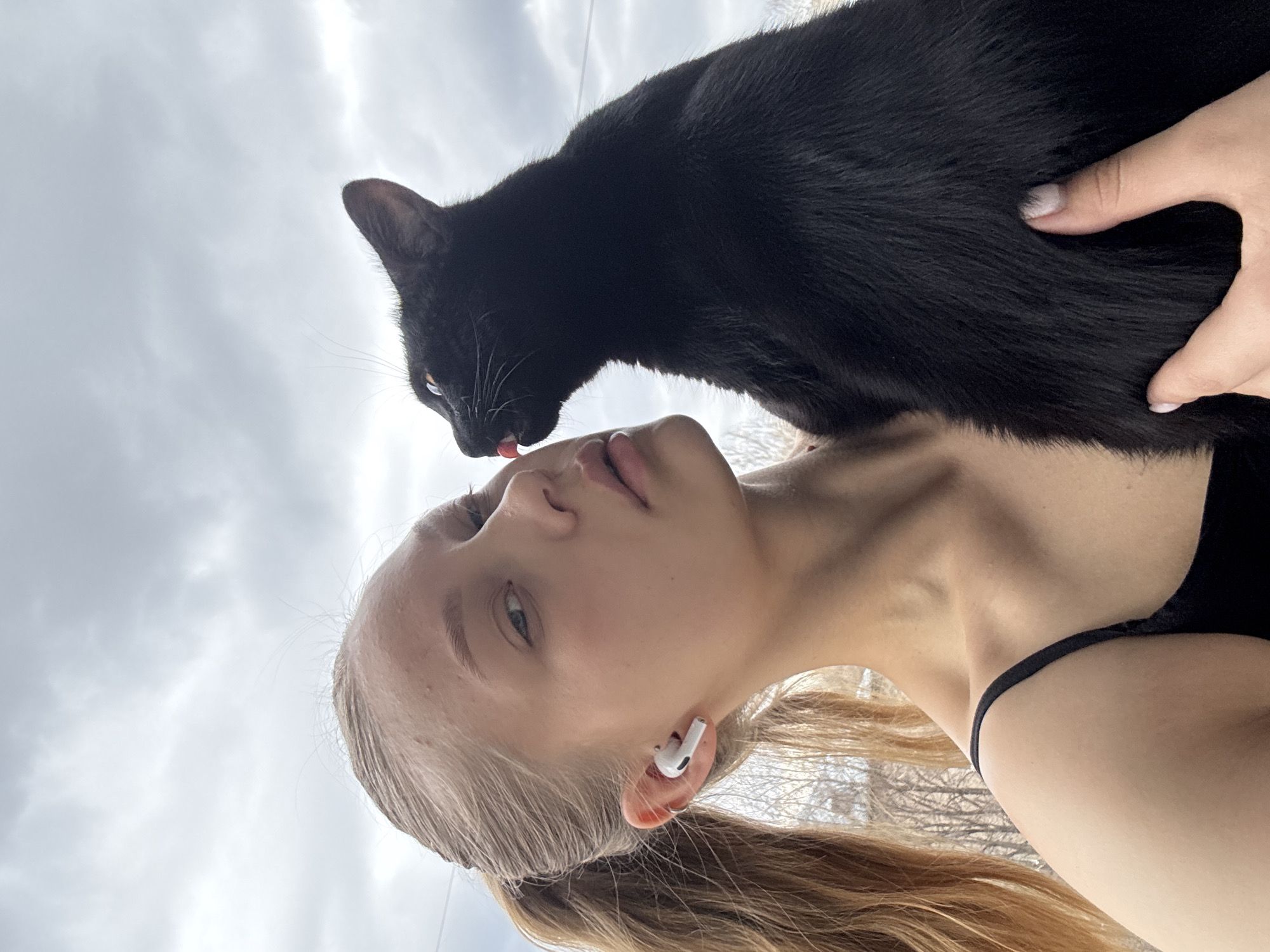 A woman with blonde hair and an AirPod in her ear is looking at the camera. A black cat is on her shoulder licking her lip. The sky is overcast. The location is likely Chaikovsky, Russia.