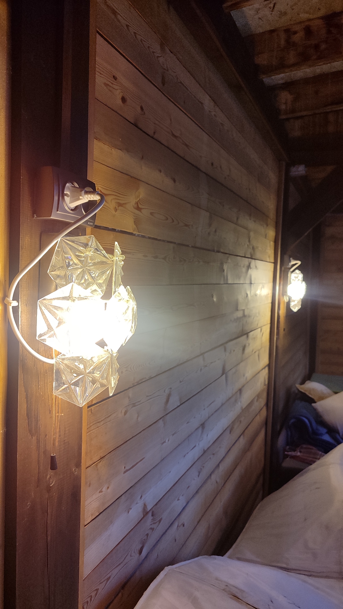 In an interior room with wooden plank walls and ceiling, an illuminated plug is attached to an outlet on the left. A white cord extends from the plug to a wall lamp with a white curved arm and a decorative glass lampshade composed of geometric, faceted shapes. The lampshade is lit by a bright bulb, casting a warm glow on the surrounding wood. Further to the right, another similar wall lamp is visible, also illuminated. In the background on the right, a bed with white bedding and pillows is partially visible, along with some folded blankets or clothing. The scene appears to be a dimly lit bedroom or living space, possibly a cabin or a room with a rustic aesthetic. The exact location within Mirny, Russia is not specified by any visual cues in the image.