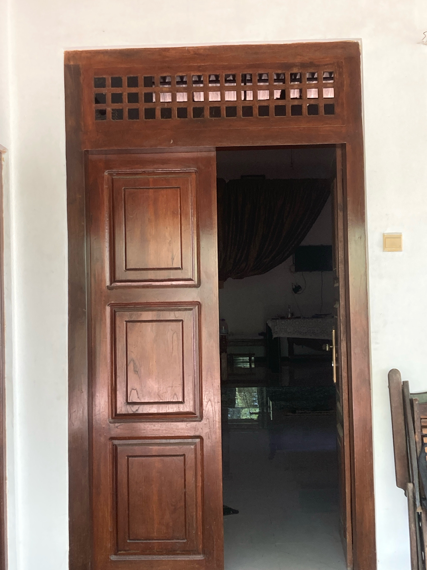 The image shows a wooden door with a ventilation grate at the top, which is ajar and reveals a dimly lit interior. The door itself has three recessed panels and is made of dark wood. To the right of the door, a portion of a white wall is visible, with a light yellow electrical outlet. Further to the right, part of a wooden chair is visible. Inside the room, a dark curtain hangs, and a television is mounted on the wall. A table with a patterned cloth is also present. The environment suggests a residential setting in Daladagama, Sri Lanka.