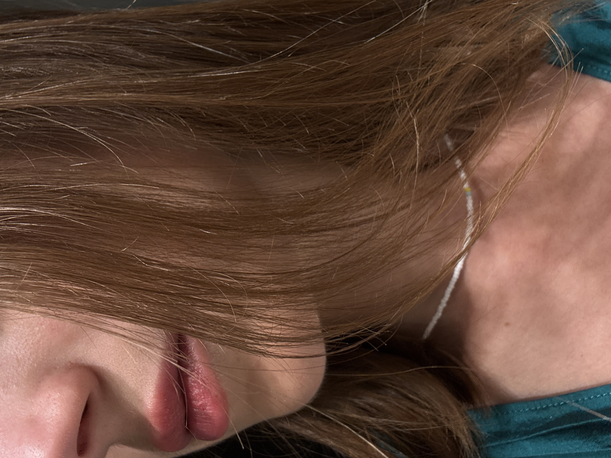 The image is a close-up of a person's face and upper body, focusing on the mouth, nose, and hair. The person has fair skin and light brown hair, which is styled to fall around their face. They are wearing a teal-colored garment and a delicate beaded necklace. The background is dark and out of focus. There are no discernible landmarks or environmental cues to suggest a specific location within Bečići, Montenegro.