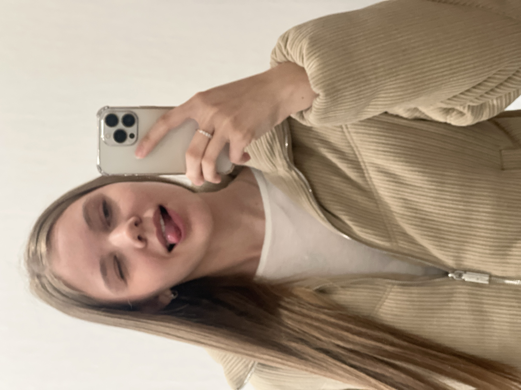 A young woman is captured in a playful selfie, with the image rotated sideways. She has long, straight light brown hair and is seen from the chest up. She wears a beige, ribbed zip-up jacket over a white t-shirt. In her right hand, she holds a smartphone, which appears to be an iPhone with a three-camera system, enclosed in a clear case. A delicate ring with multiple small, light-colored beads is visible on her left ring finger. Her expression is lighthearted; she is winking with one eye and sticking out her tongue. The background is a plain, light-colored wall, suggesting an indoor setting. No specific time of day or explicit location cues are visible within the image content, though the location context is Votkinsk, Russia. There is no visible text.