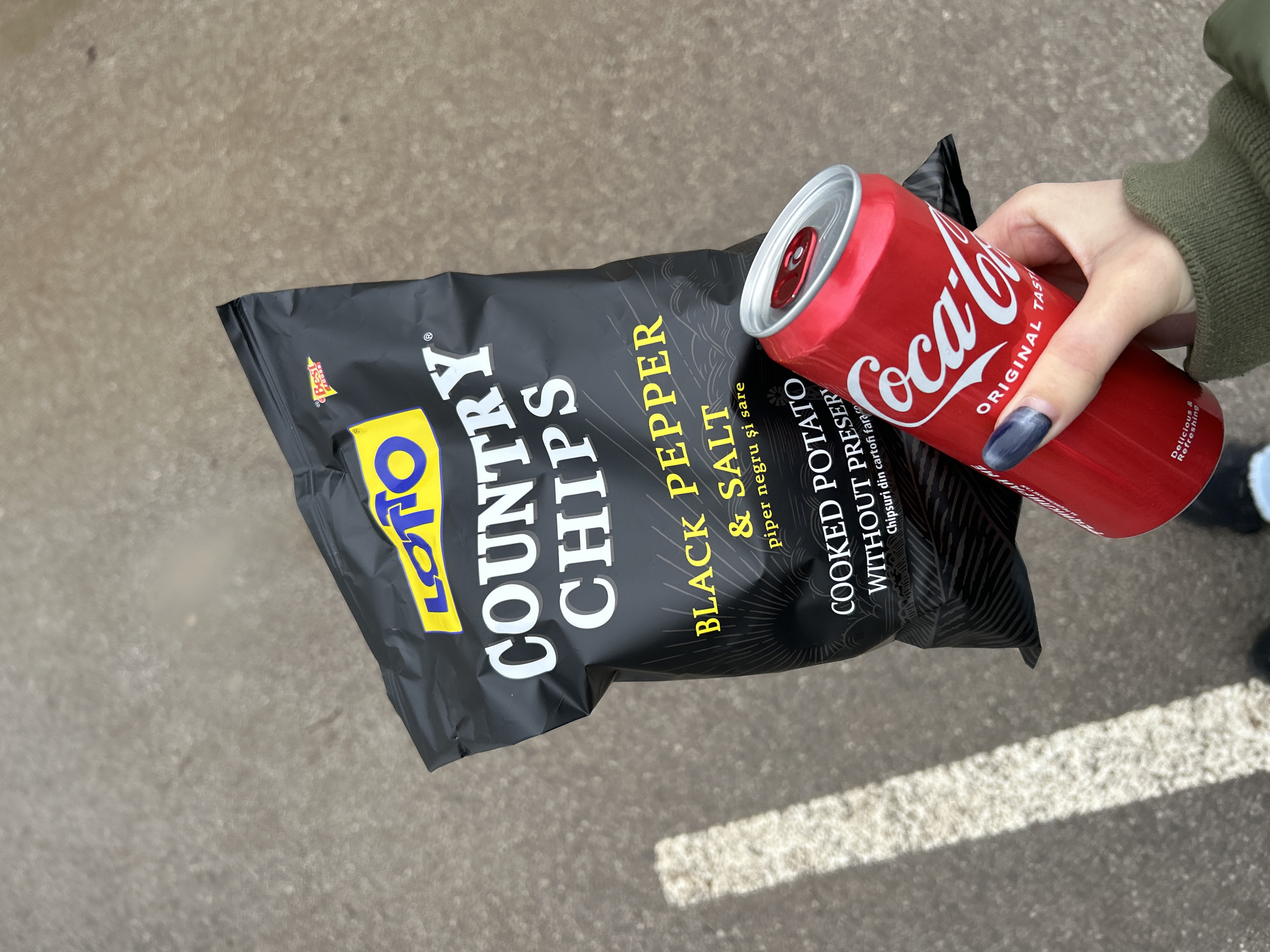 The photograph shows a hand holding a red Coca-Cola can and a black bag of Lotto Country Chips Black Pepper & Salt. The background is a gray asphalt surface with a white painted line visible in the lower right corner. The chips are labeled as "COOKED POTATO WITHOUT PRESERVATIVES." The brand logo "BEST FOODS" is visible on the top left corner of the chip bag. The scene appears to be outside on a cloudy day. The exact location is not discernible from the photograph.