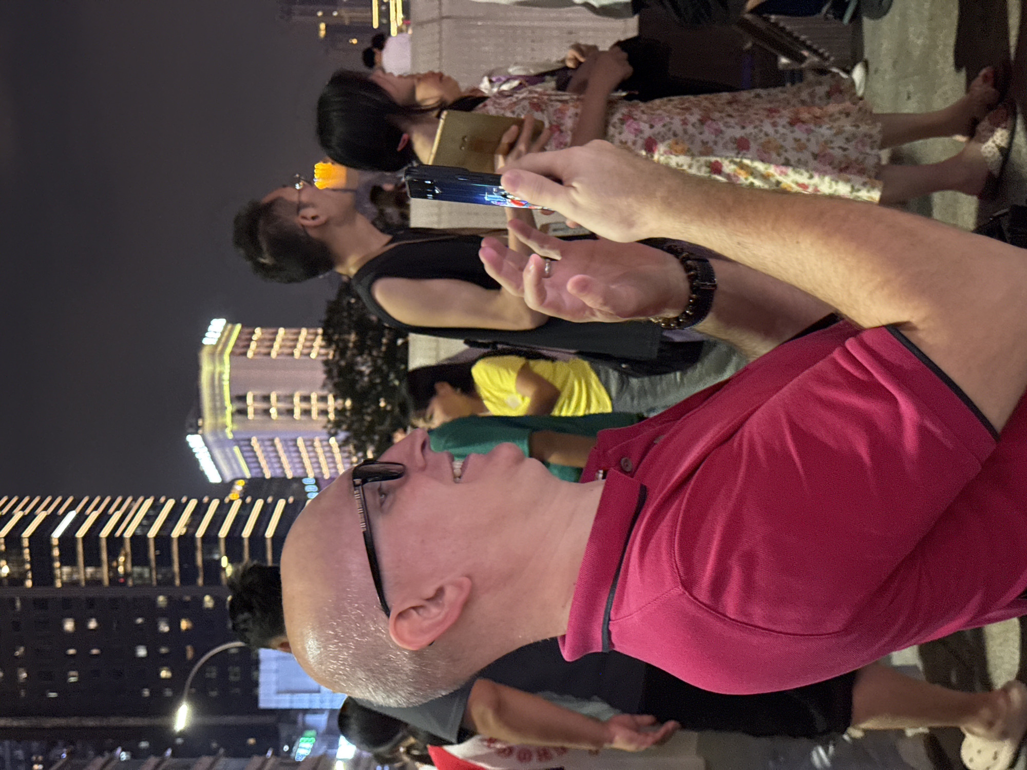 In Yuzhong District, China, a bald Caucasian man wearing glasses and a pink polo shirt is captured from the chest up, smiling and looking to his right while holding a smartphone and a light-colored object in his hands. The man's right hand is extended, holding the phone which displays a colorful image. The background features a blurred cityscape at night, with tall buildings illuminated by bright lights. A woman in a floral dress and another person in a black sleeveless shirt are partially visible to his right. The atmosphere suggests a public gathering or event.