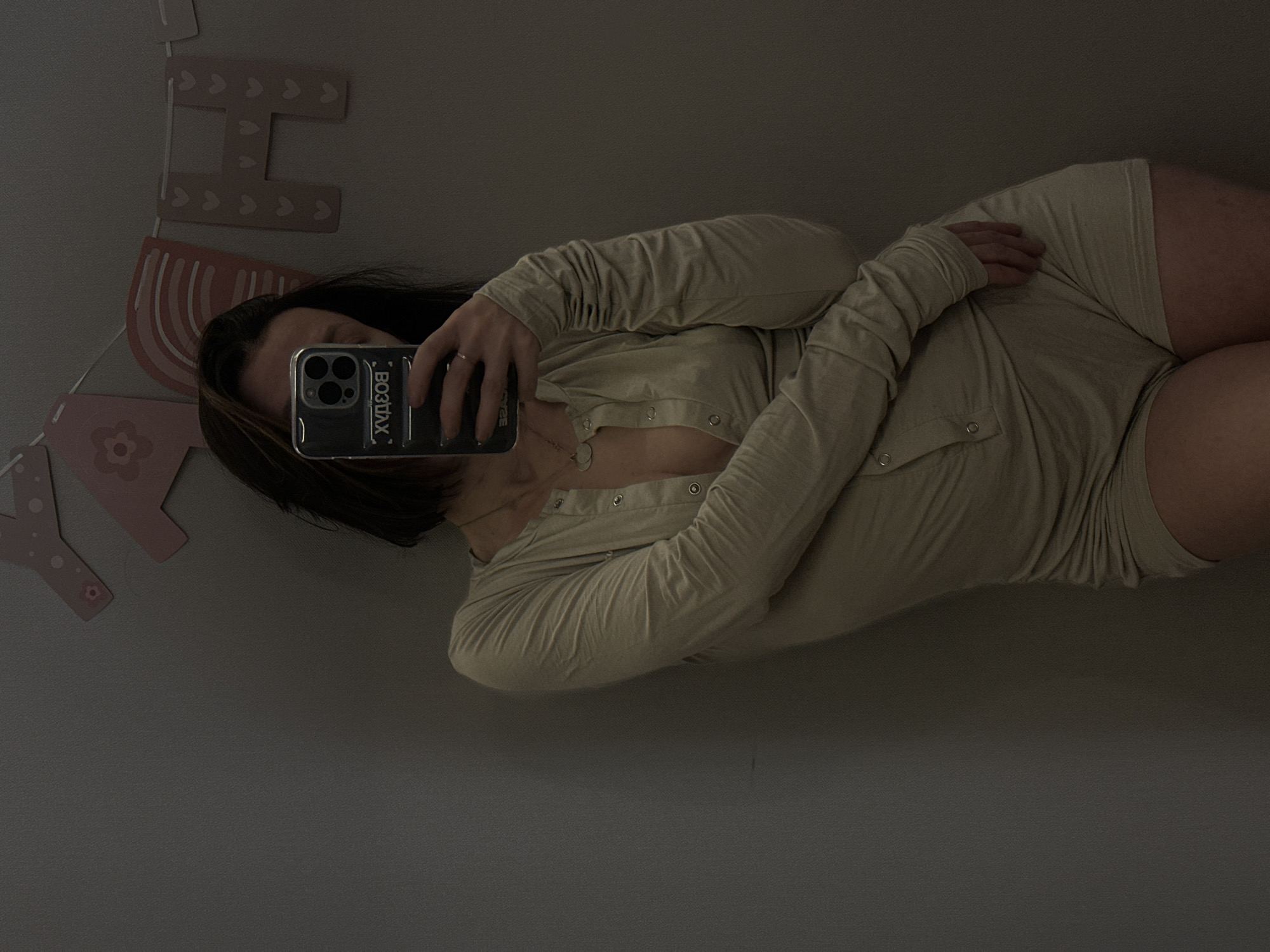 A person is taking a selfie with a smartphone. They are wearing a light-colored, long-sleeved garment with buttons down the front. The phone case has the word "BOTTAX" printed on it. The background includes a wall decoration with letters and a rainbow design. The image does not provide any indication of the city or location in Votkinsk, Russia.
