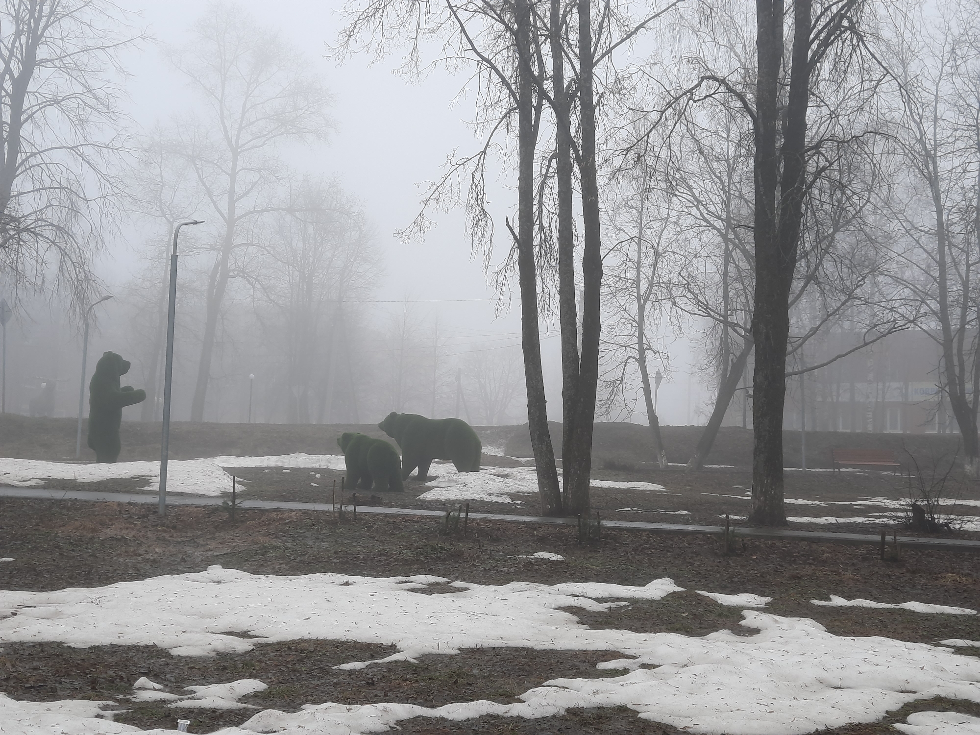 The image shows a foggy scene in what appears to be a park or open space. Patches of snow cover the ground, mixed with bare earth and vegetation. Several leafless trees are visible, their branches creating a stark contrast against the heavy fog. In the background, one can discern a building and a bench, suggesting a park or recreational area. In the midground, there are three green bear-shaped topiary bushes. The photograph was taken in Krasnogorskoye, Russia.