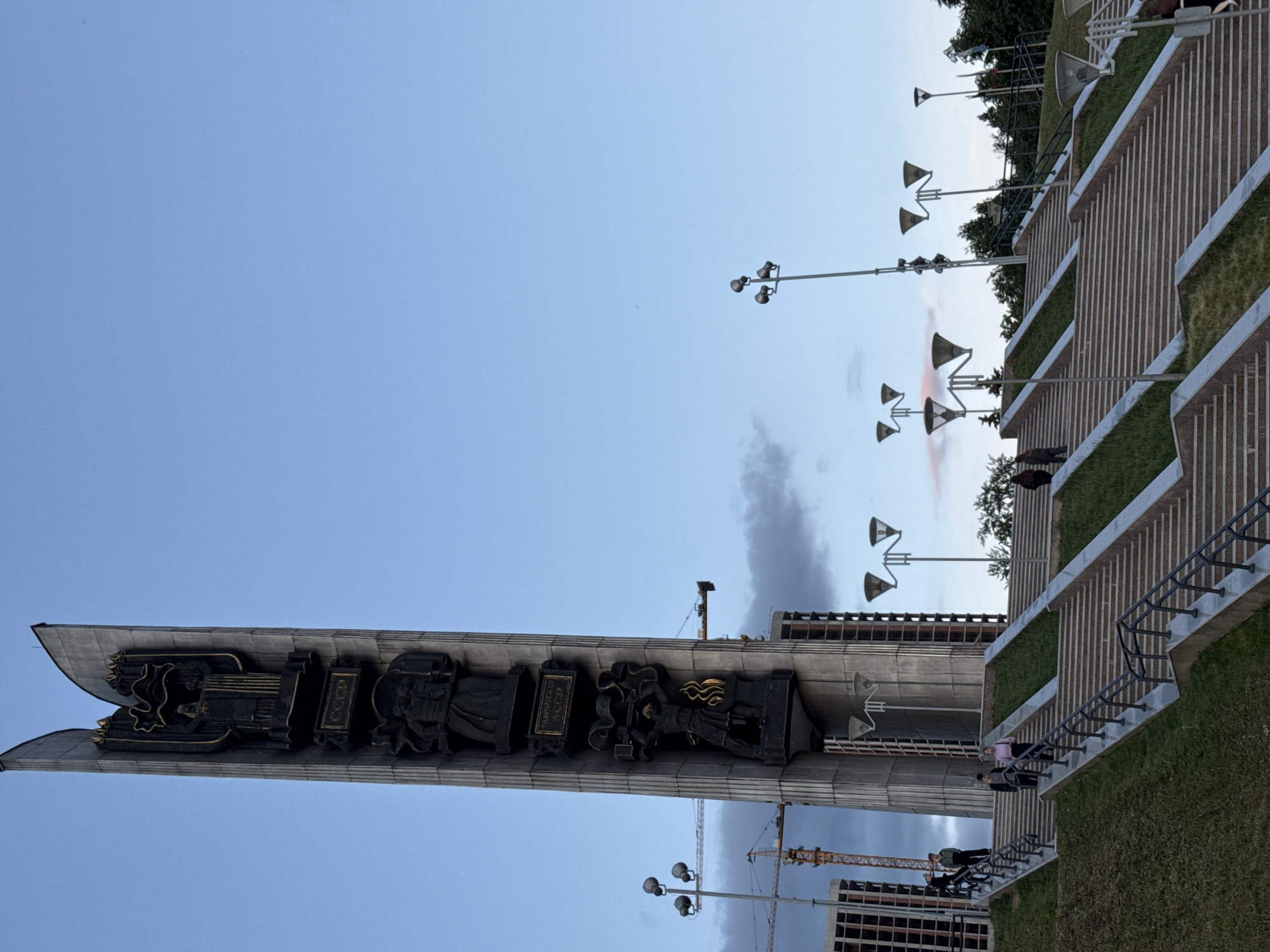 The image displays the Monument of Friendship of Peoples in Izhevsk, Russia, under a bright blue sky with scattered clouds, indicating daytime. The monument, a tall, imposing concrete or stone structure, features prominent dark bas-reliefs along its surface. These reliefs depict various stylized figures and symbols, and notably include the Cyrillic text "Удмуртская АССР" (Udmurt ASSR) and "СССР" (USSR), signifying its Soviet-era origins and connection to the Udmurt Autonomous Soviet Socialist Republic.
To the right of the monument, a series of broad, terraced stairs and walkways ascend, interspersed with sections of green grass. Several multi-lamp streetlights are positioned across these terraces and in the foreground. A few individuals are visible on the stairs and walkways, casually moving or standing, suggesting general public access to the area. In the background, particularly behind the monument's base and beyond the terraced landscape, glimpses of modern buildings are seen, including one under construction with a large yellow crane beside it. Trees and other vegetation are present towards the upper right of the terraced area. The scene depicts a public commemorative space, with people present but no specific event taking place.