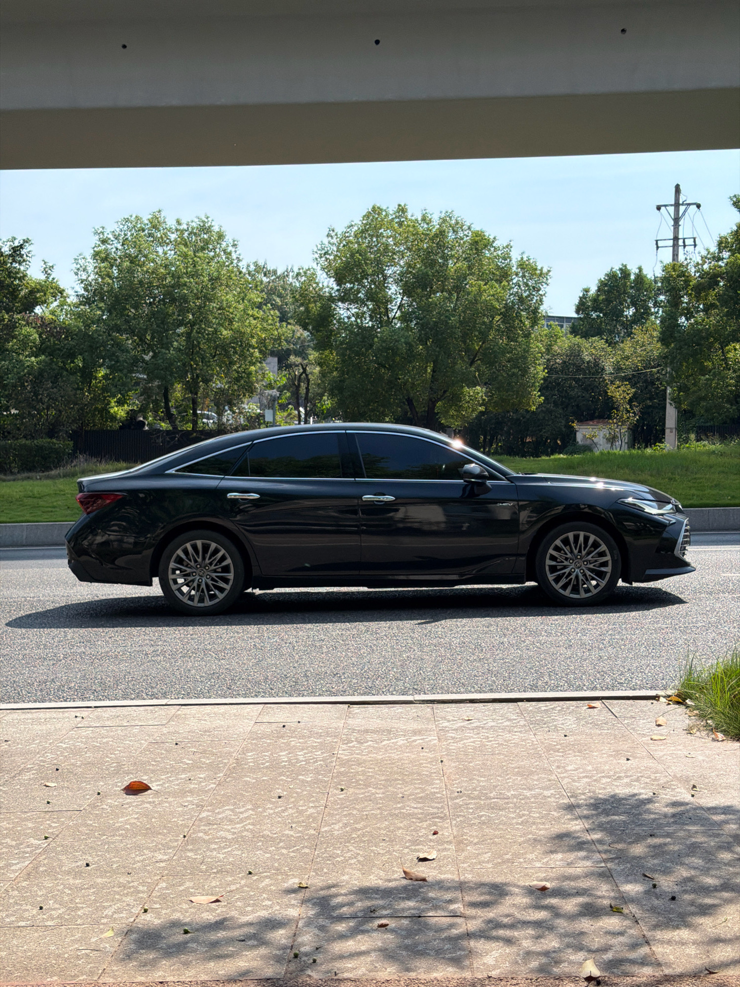 A black sedan is parked on a road in the city of Jiangjin, China. The car is viewed from the side, with the front of the vehicle facing towards the right of the frame. The sedan is a modern model, featuring tinted windows and alloy wheels. The road surface is asphalt, with a visible painted white line marking the edge of the lane. Adjacent to the road is a grassy verge and a sidewalk paved with large rectangular tiles. Fallen leaves and small dark objects are scattered across the sidewalk. Beyond the greenery, a utility pole with wires is visible against a clear blue sky. The scene appears to be captured during daylight, with shadows cast from an overhead structure and trees.