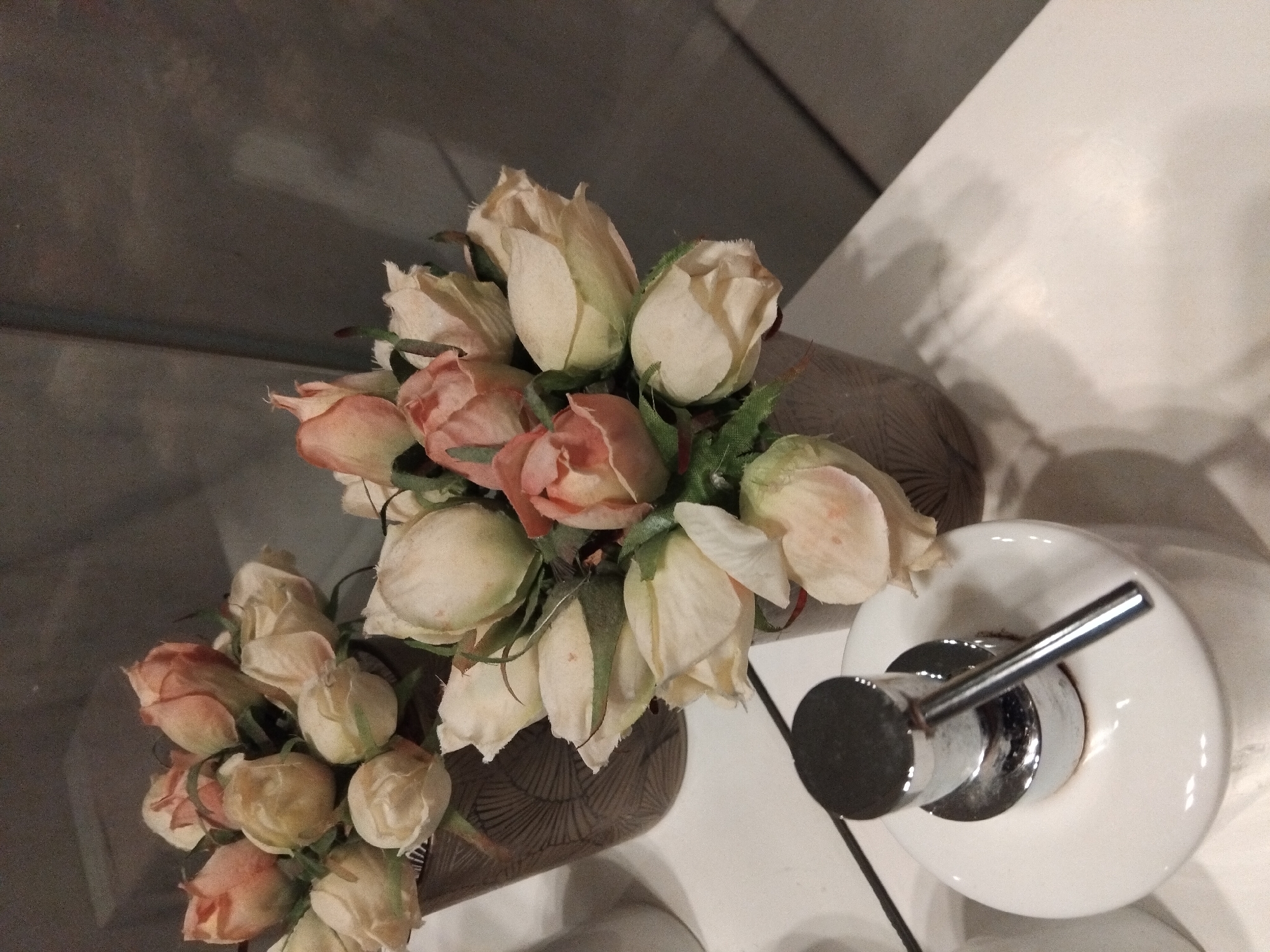 A top-down, slightly angled close-up shot shows a bouquet of artificial roses in muted pinks and creams arranged in a woven brown vase. The roses are depicted with tightly furled petals, and some show hints of green leaves and stems. To the right of the vase, a white ceramic soap dispenser with a silver pump mechanism is visible. The dispenser is round and sits on a white surface. The background is a dark, textured gray, possibly a wall or countertop, with a diagonal white surface on the right, creating a shadow effect. The lighting is dim, suggesting an indoor setting, possibly a bathroom or vanity area, with some reflections on the dispenser. No people are present in the image. The image appears to be taken in low light conditions. There is no visible text.