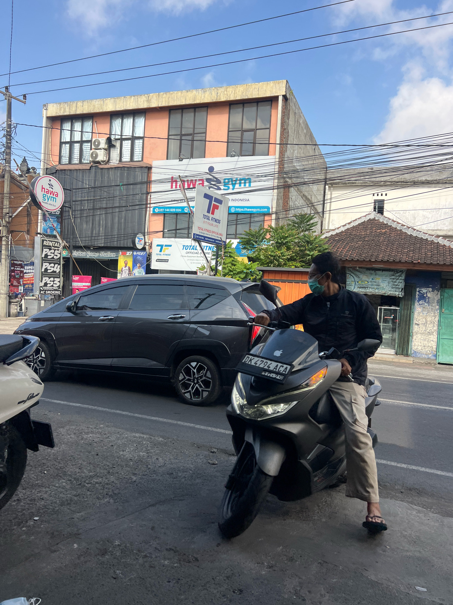 In Denpasar, Indonesia, a man wearing a light blue face mask and a dark long-sleeved shirt sits on a dark gray motorcycle, facing right. He is wearing light-colored cargo pants and black flip-flops. The motorcycle has a license plate that reads "DK 4794 ACA". Behind the motorcycle, a dark gray SUV is parked on the street, partially obscuring the motorcycle's front wheel. The street is paved with asphalt and appears to be a busy urban environment. In the background, several buildings are visible, including one with an orange facade and large windows. Signs for "Hawa Gym" and "Total Fitness" are prominently displayed on the buildings. Power lines and utility poles crisscross the sky. The overall scene depicts a typical street view in a bustling Indonesian city.