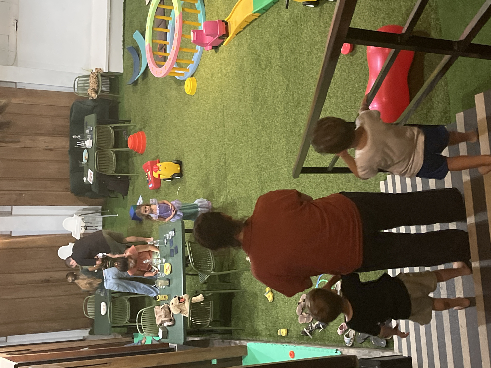 The image depicts an indoor play area in North Kuta, Indonesia. The space is covered with artificial turf and features several tables and chairs, a sofa, and a variety of children's toys and play equipment, including a toy car, a pink ride-on toy, a large red inflatable, and a colorful play structure. Several adults and children are present. In the foreground, two young boys are descending a striped staircase, with an adult woman standing next to them. Further back, a group of adults and children are gathered around a table, with some interacting and others seated. The overall atmosphere suggests a casual gathering or a children's playdate.