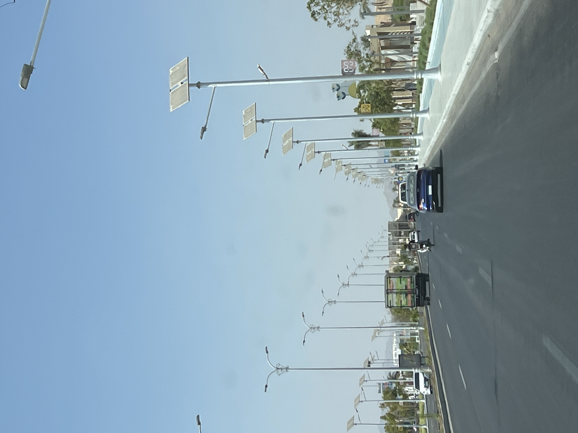 The image depicts a wide, paved road in Sharm El Sheikh, Egypt, under a clear blue sky. The road features multiple lanes with white dashed lane markers. Along the right side of the road, there is a well-maintained sidewalk or pedestrian path, bordered by a curb. A consistent line of modern streetlights, many equipped with solar panels, are positioned along the sidewalk, extending into the distance.
Several vehicles are visible on the road. In the foreground, a blue sedan is traveling away from the camera. Further ahead, a white truck and a motorcycle are also moving in the same direction. A white car is parked on the left side of the road. In the background, there are buildings and landscaping, including palm trees and manicured greenery, suggesting an urban or resort area. The architecture appears modern. The overall scene conveys a sense of a well-developed and functional urban environment.