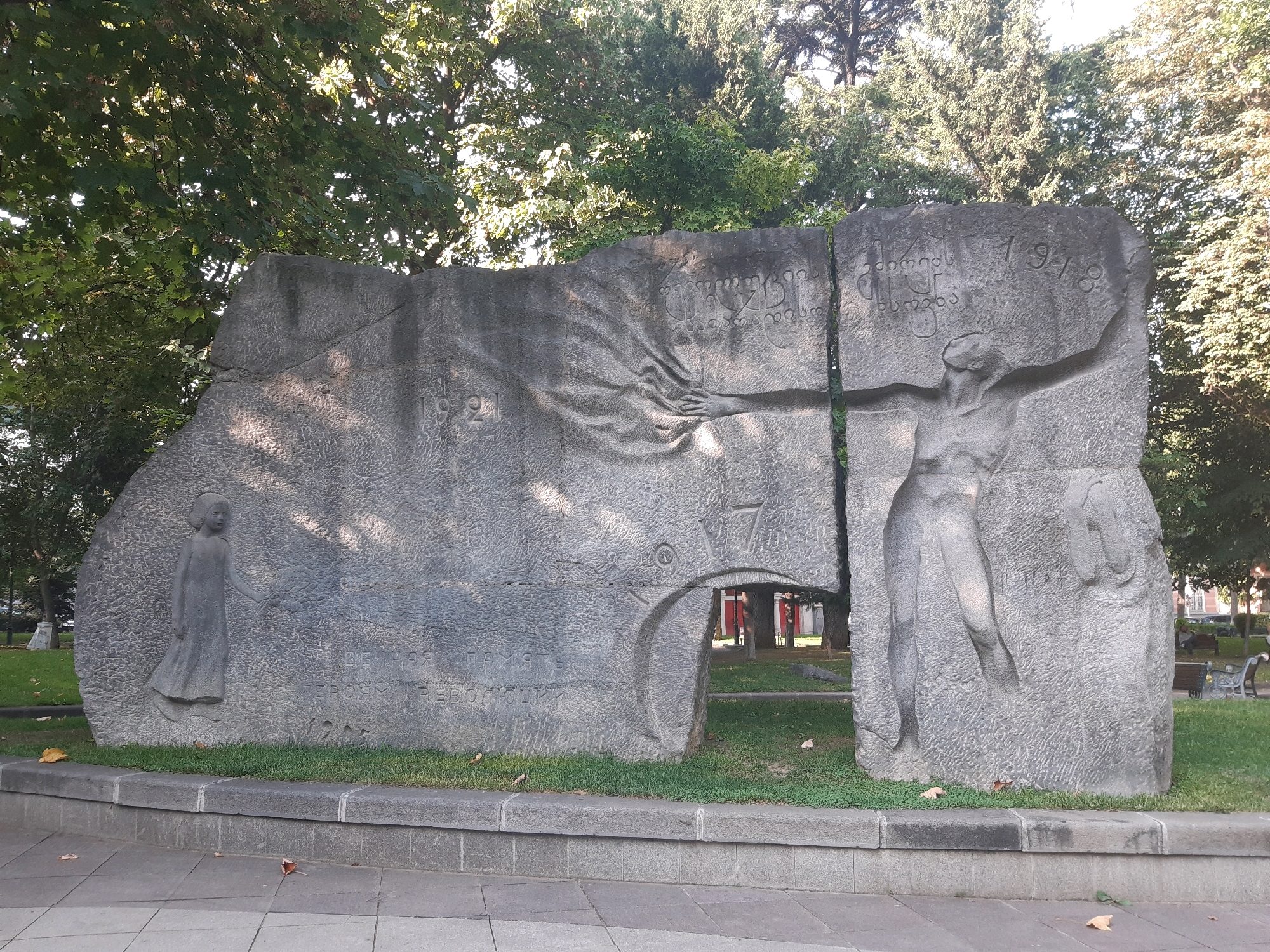 The image shows a large stone monument in a park in Tbilisi, Georgia. The monument is abstract in design and features carved figures and text. On the left side of the monument, there is a carving of a young girl holding a bouquet of leaves. To the right of the girl, there is a large, cracked stone structure with carvings of human figures and Georgian script. One prominent carving on the right depicts a woman with outstretched arms, seemingly in a gesture of grief or defiance, with the year "1918" carved above her. The monument is set against a backdrop of lush green trees, with sunlight filtering through the leaves, casting shadows on the stone. In the background, there are benches, more trees, and a hint of urban structures, suggesting it is located in a public space within the city. The ground in front of the monument is paved, with a low stone border separating it from the grass. A few fallen leaves are scattered on the grass and pavement.