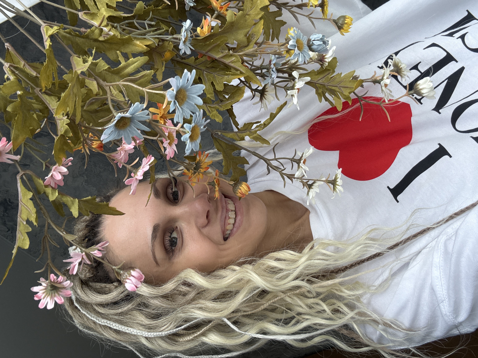 A person with blonde, wavy hair interspersed with white and brown strands lies on a surface. The person is wearing a white t-shirt with a design, including a large red heart. A bouquet of artificial flowers with blue, pink, white, and yellow blossoms and green leaves is positioned around their head. The surface beneath appears to be gray. The location is in an unknown city.