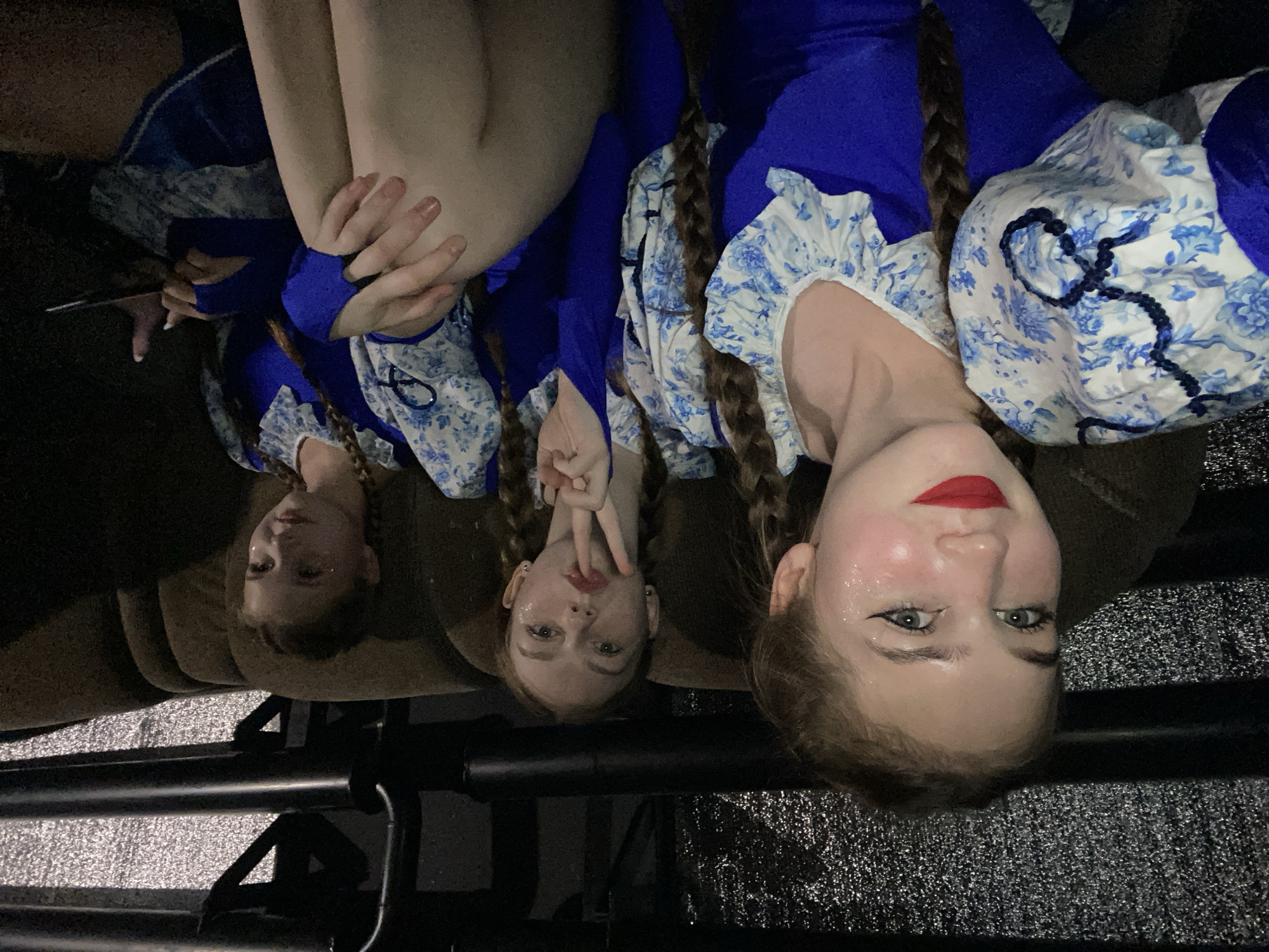 Three young females are seated on a brown couch in what appears to be a backstage or dressing room area. They are all wearing similar blue and white floral costumes with voluminous sleeves. The female on the left is in the foreground, looking directly at the camera with bright red lipstick and glitter on her cheeks and forehead. Her hair is in a braid. The female in the middle is also looking at the camera, holding up two fingers in a peace sign and has glitter on her cheeks. Her hair is in braids. The female on the right is looking away from the camera, also with glitter on her cheeks, and is holding a dark object, possibly a phone, in her right hand. The environment is dimly lit, with a textured metallic wall and black pipes visible in the background. There is no direct indication of the city of Chaikovsky, Russia, within the image itself.