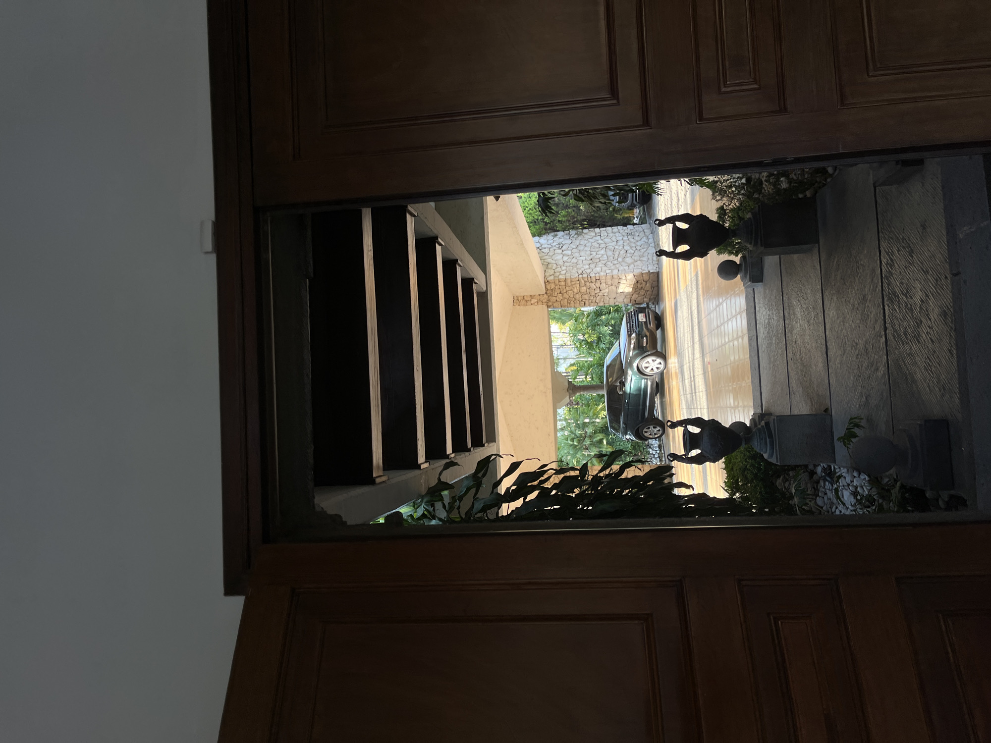 A view through a partially open dark brown wooden door reveals an outdoor scene with a black car parked on a paved surface. Two urns, adorned with statues, are positioned on either side of the doorway. The location appears to be San Bernardino Tlaxcalancingo, Mexico. The scene is characterized by a mix of natural elements and architectural features.