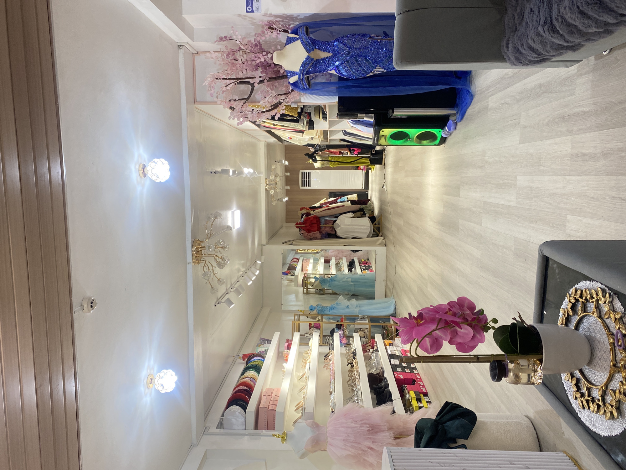 The image shows the interior of a retail store that appears to specialize in formal wear and accessories. The store is brightly lit with multiple ceiling lights. On the left side of the store, there are white shelves displaying rows of shoes and hats. A pink tulle dress is displayed on a mannequin in the foreground. In the center of the store, there are more shelves with shoes and boxes. Further back, mannequins are dressed in elaborate gowns, one in blue and another in light blue. There are racks of clothing and a decorative pink tree with blossoms. The floor is made of light-colored wood planks. In the foreground, a table holds a potted orchid and a small glass bottle. A gray sofa with a furry throw is visible on the right side. The store is located in Davao City, Philippines.