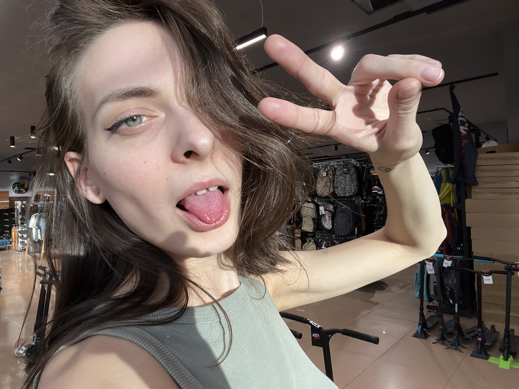 This close-up selfie captures a young woman inside a retail store, likely located in Krasnoyarsk, Russia. She is the central subject, positioned with her face close to the camera. She has long, dark brown hair, which partially obscures the right side of her face. Her eyes are a light green, accentuated by prominent black winged eyeliner. She is sticking out her tongue in a playful expression and holds her left hand up, making a "peace" or "victory" sign with her index and middle fingers extended. She wears a light green or mint-colored sleeveless top.
The background shows a brightly lit indoor retail environment. Displays with various merchandise, including what appear to be backpacks or bags, are visible on racks. Towards the lower right, several scooters or similar wheeled sports equipment are arranged on the floor. Overhead, modern strip lighting and circular light fixtures illuminate the space.
Notable details include a small, circular tattoo on her left wrist, featuring an abstract symbol. Faint text reading "KICK" is visible on the handlebar of a scooter in the lower right of the image. The overall impression is one of playful self-expression within a casual shopping setting during the daytime.