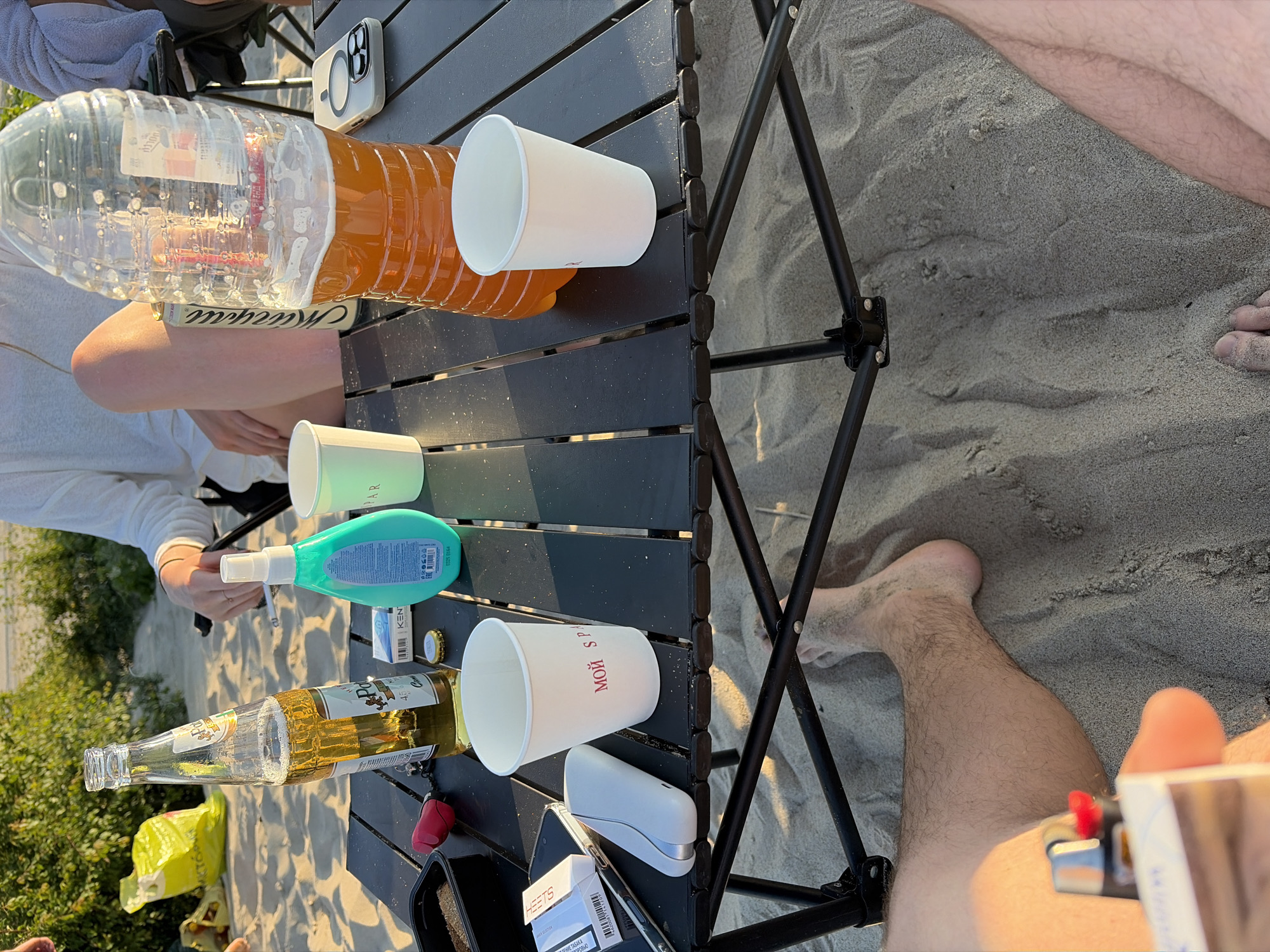 This is a medium shot, eye-level, outdoor photograph taken in the city of Povarovka, Russia. The scene is on a sandy beach during daytime, with lush green foliage visible in the background. A foldable black table is set up on the sand, displaying various items.
On the table, there is a tall, clear plastic bottle partially filled with an amber liquid, possibly beer or a soft drink. Next to it is a glass bottle of beer, with condensation on its surface and some foam at the top. There are two white paper cups, one with red Cyrillic text that appears to read "мой спорт" (my sport). A turquoise colored bottle of what looks like hand sanitizer or soap is also present. To the left, a pack of cigarettes labeled "HEETS" and a phone are on the table, along with a small black container filled with a granular substance. A red object, possibly a lighter or a tool, is also visible.
In the foreground, a person's bare legs and feet are partially visible, covered in sand. The sand appears coarse and light in color. The overall atmosphere suggests a relaxed outdoor gathering.
