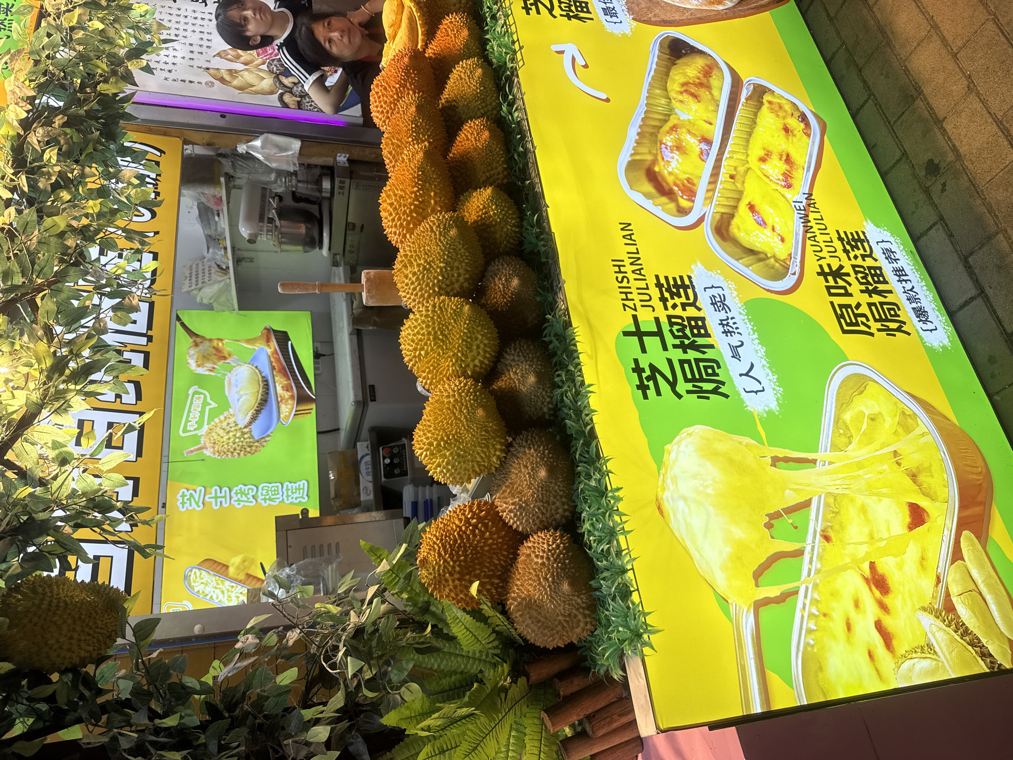 This image depicts a street food stall, likely in Qinhuangdao, China, specializing in durian-based desserts. The stall features a bright yellow banner with green accents, displaying images of durian fruit and baked durian dishes topped with melted cheese. Several whole durians are prominently displayed on a bed of artificial grass, creating a visually appealing setup. In the background, a commercial mixer and other kitchen equipment are visible, suggesting the food is prepared on-site. Two women are present, one in the foreground looking towards the camera, and another in the background glancing away. The overall scene suggests a bustling, outdoor food vendor environment, indicative of a marketplace or busy street.