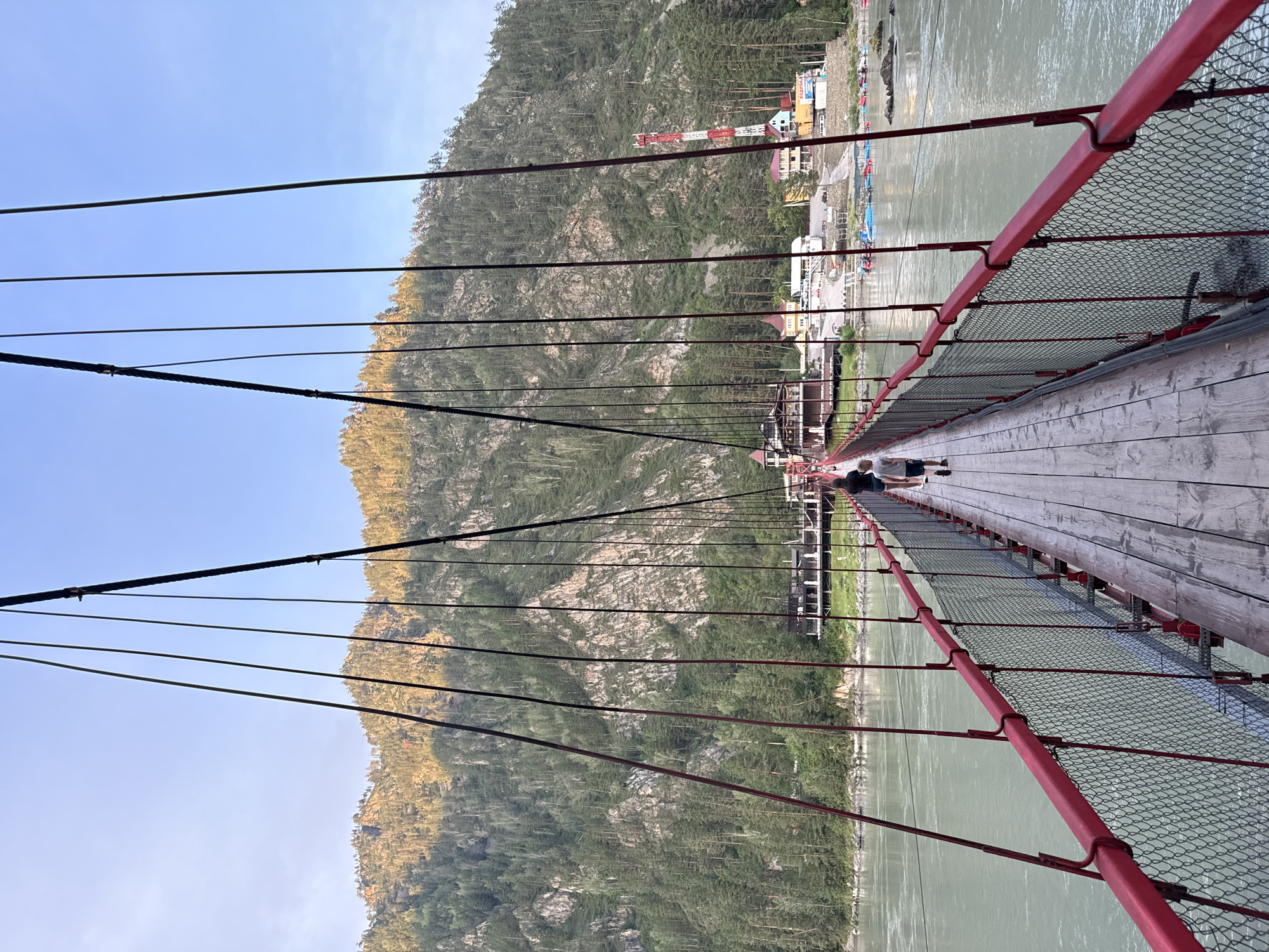 The image captures a pedestrian suspension bridge over a wide, calm river in Barangol, Russia. The bridge has red railings and a wooden walkway. Two people, an adult and a child, are walking away from the camera across the bridge. On the far bank of the river, there is a cluster of buildings, including a large hotel or resort with red roofs, and a tall red and white tower that appears to be a telecommunications tower. The background is dominated by a steep, densely forested mountain with rocky outcrops, bathed in the warm light of what appears to be late afternoon or early morning. Several small boats are visible near the shore on the far side. The sky is clear and blue.
