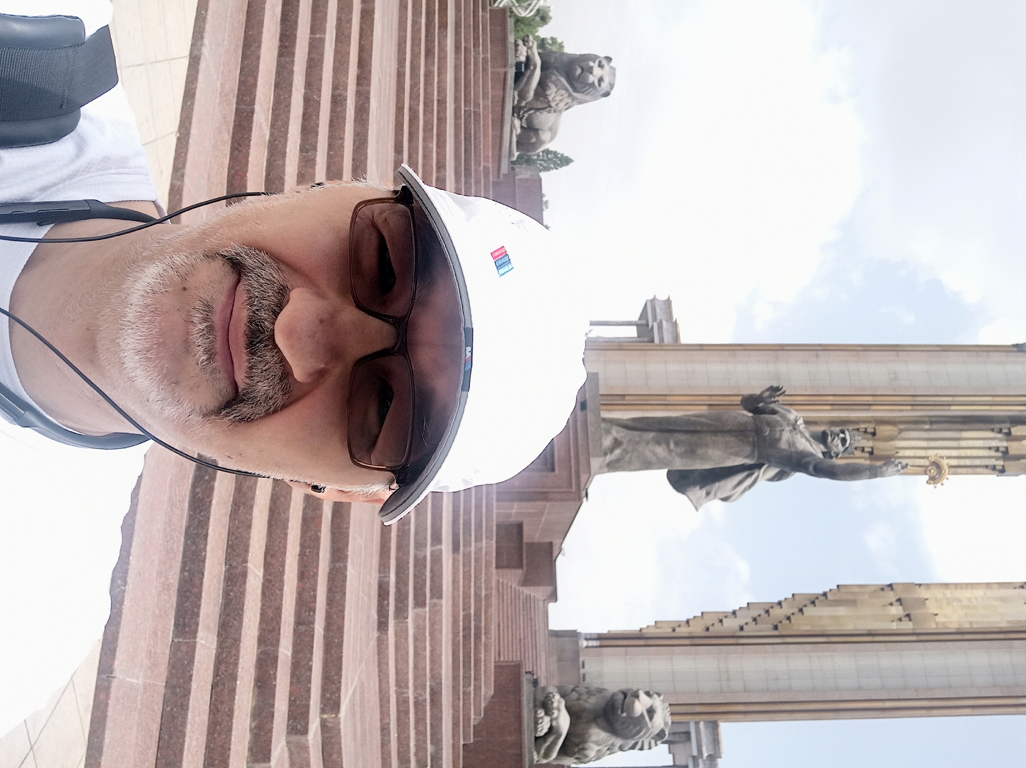 This is a selfie taken by a man in Dushanbe, Tajikistan, in front of the Rudaki Park. The man is wearing a white baseball cap and glasses, and he has a mustache and goatee. He is holding a phone to take the picture. In the background, there is a large statue of a man holding a torch, flanked by two tall pillars. On either side of the stairs leading up to the statue are two stone lion statues. The man is standing on a wide set of stairs.